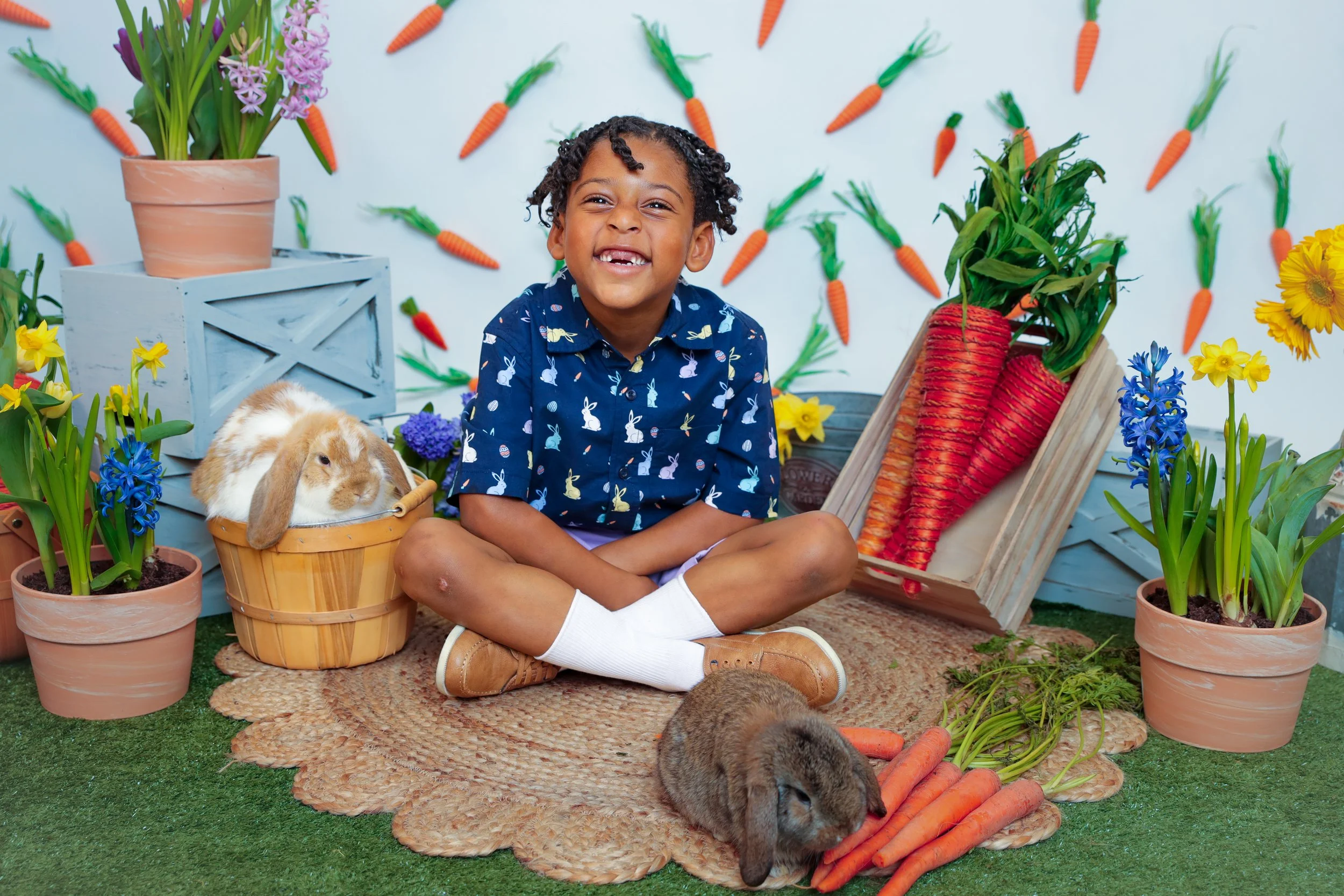 A young boy sitting cross-legged on a rug surrounded by potted flowers, carrots, and two rabbits in a colorful, garden-themed setting with a backdrop of hanging carrots and flowers.