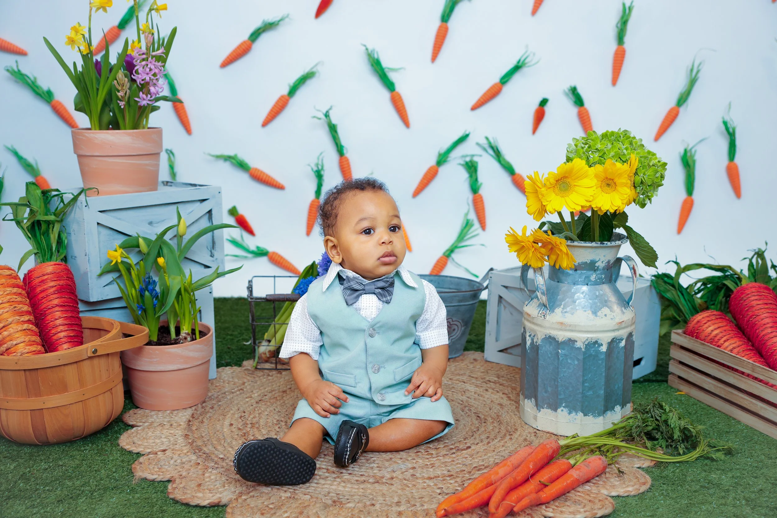 A young boy sitting on a woven rug surrounded by decorative carrots and flowers in pots, with a backdrop of hanging paper carrots