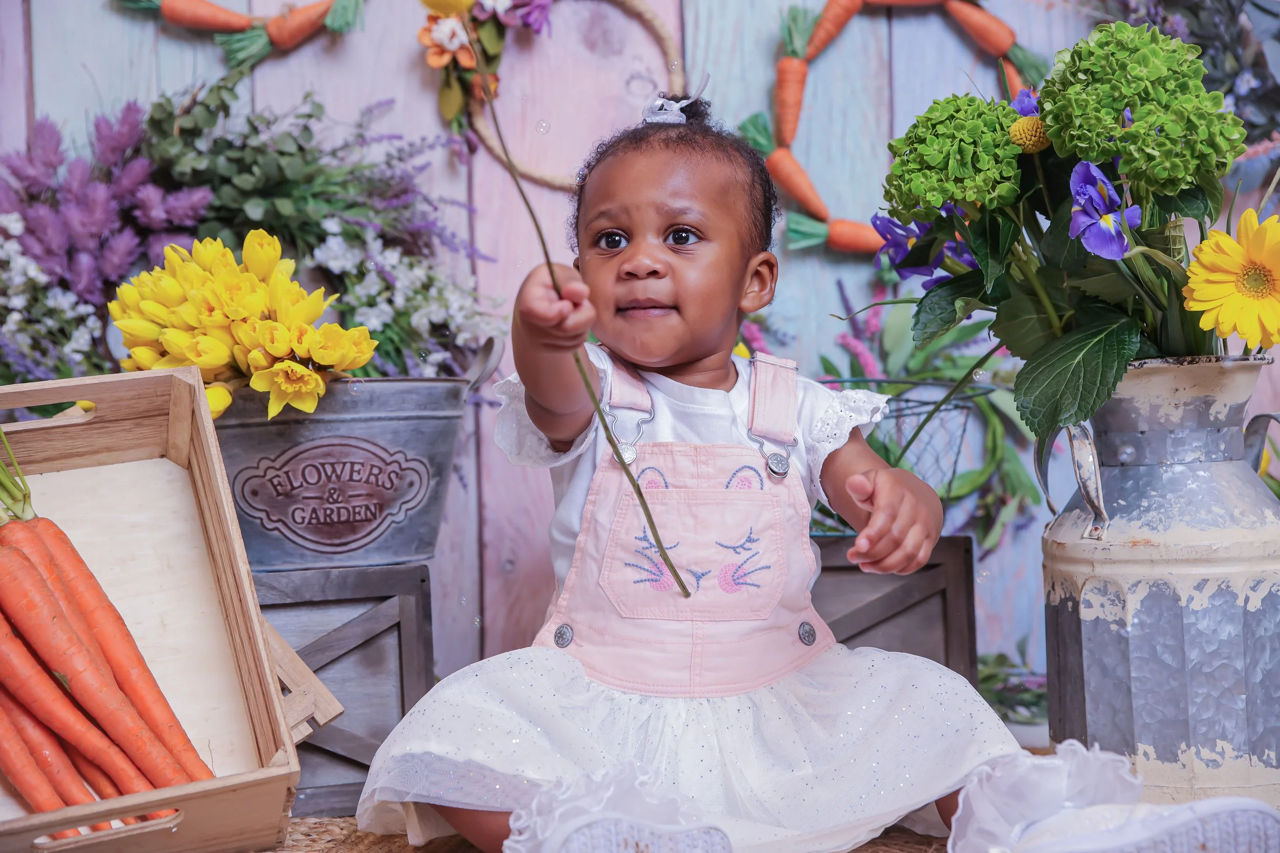 A young girl sitting among colorful flowers and vegetables, holding a thin branch, with a background of flower arrangements and garden-themed decorations.