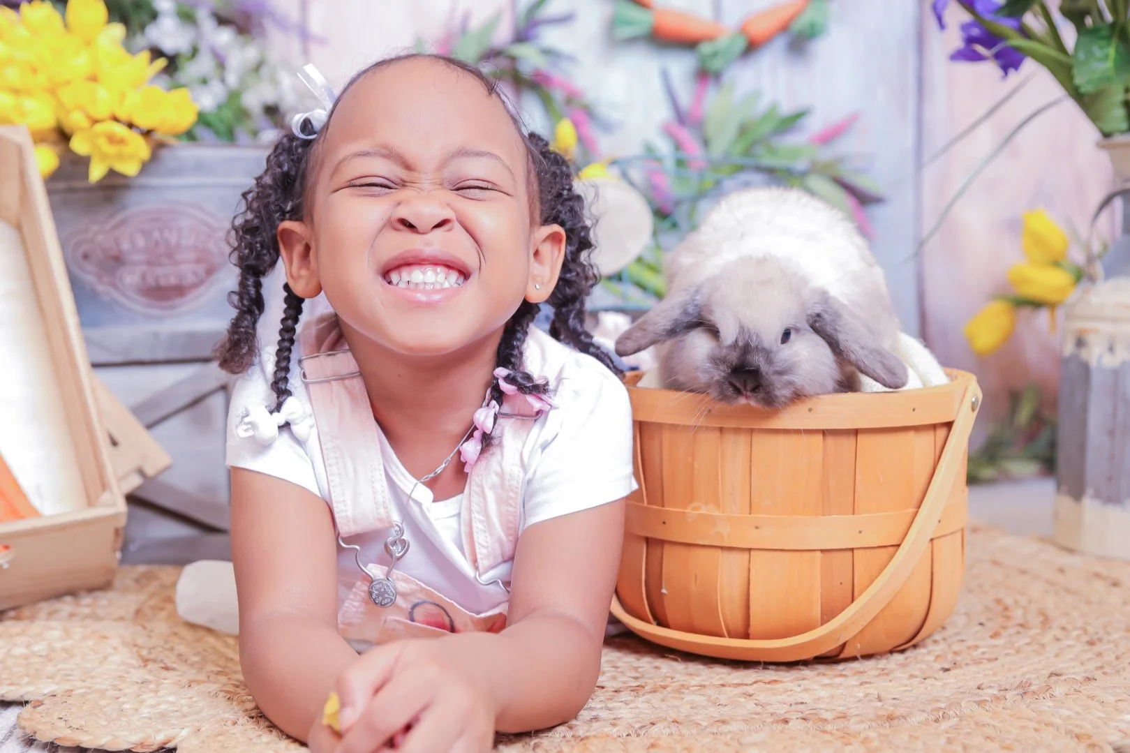 Young girl with curly hair, smiling and squinting, lying on a woven rug next to a small bunny in a wooden basket, surrounded by colorful flowers.
