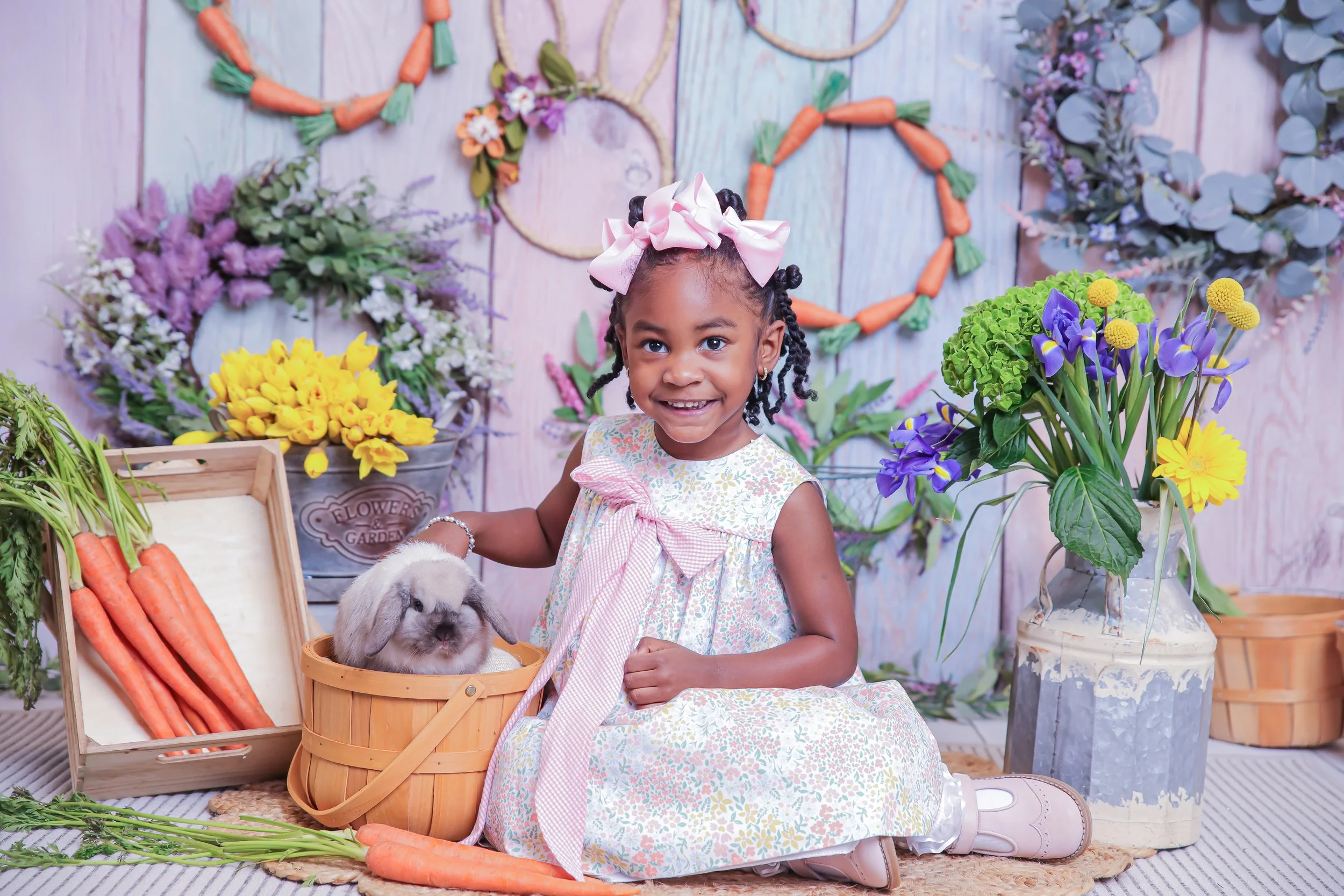 A smiling young girl with braided hair and a large pink bow, sitting on a carpet among vibrant flowers and vegetables, including carrots and a bunny in a basket, in a colorful floral setting.
