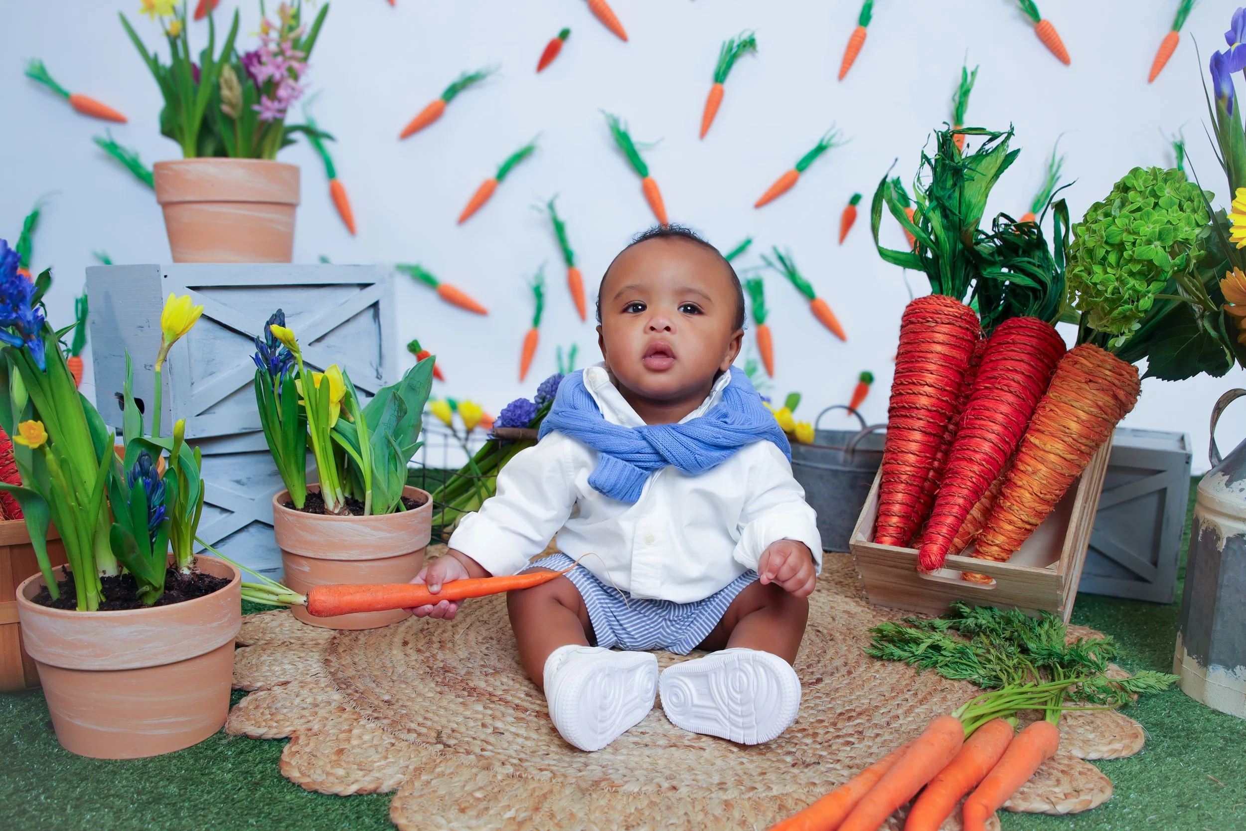 A baby sitting on a woven mat surrounded by potted flowers, carrots, and artificial carrots on a decorated backdrop.