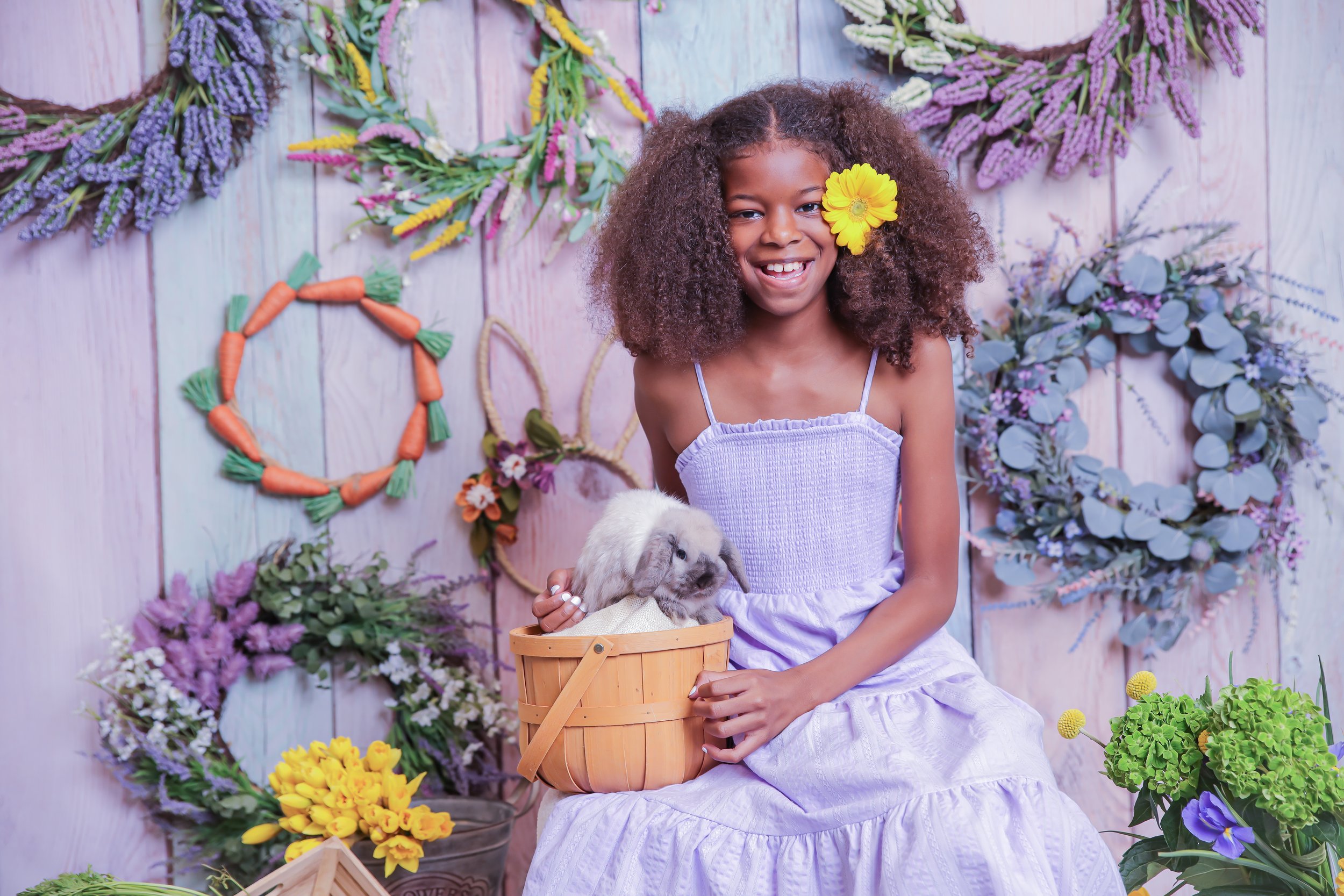 A young girl with curly hair, wearing a lavender dress and a yellow flower in her hair, sitting and smiling while holding a rabbit in a basket. The background features colorful flower wreaths and decorations.
