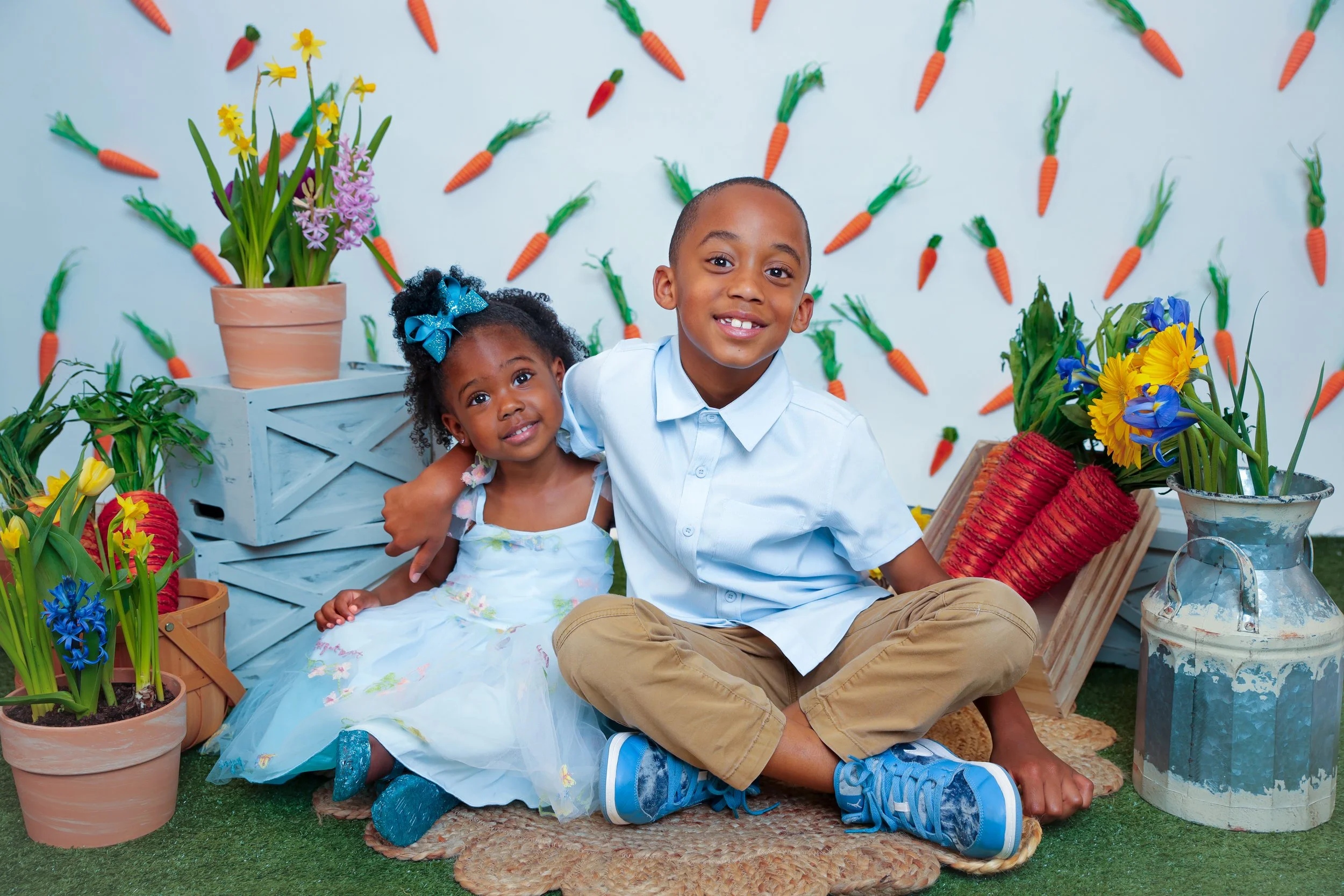 Two children sit on a mat amid colorful spring flowers, with a backdrop decorated with paper carrots on a wall.
