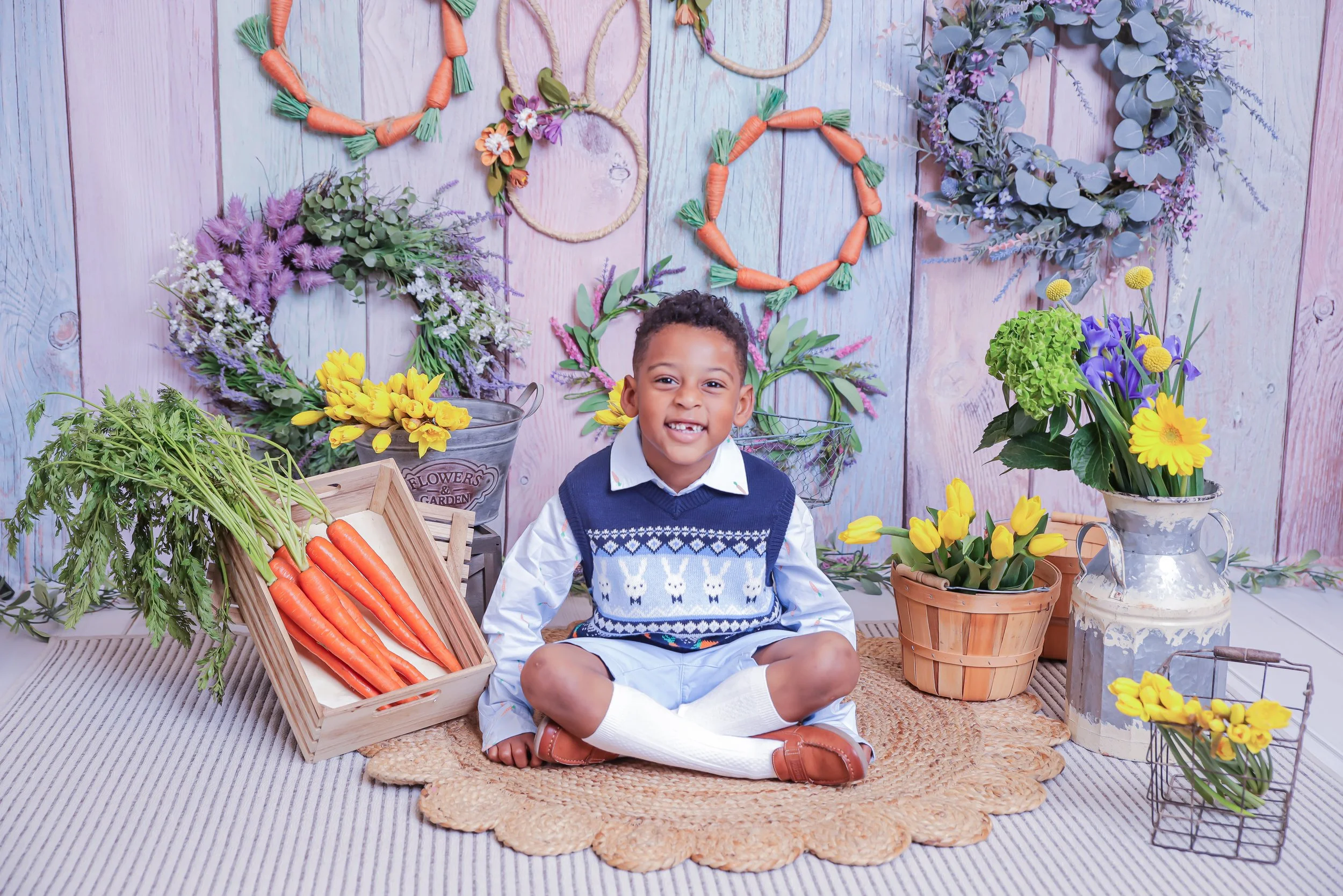 A young boy sitting cross-legged on a round woven rug surrounded by colorful flowers, wreaths, and baskets of carrots, in a decorated floral-themed studio.