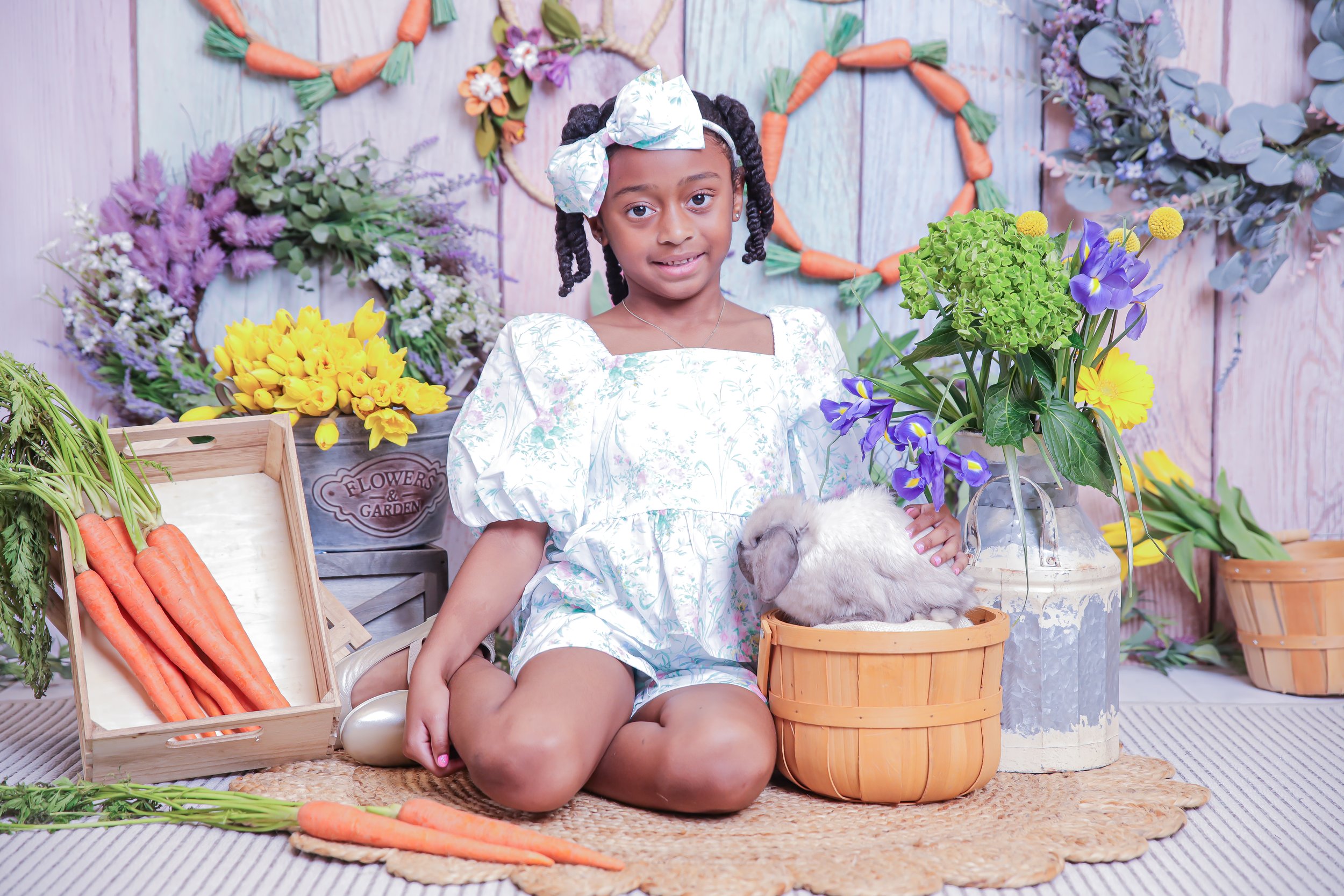 Young girl with a bow in her hair sitting among flowers and carrots, with a rabbit in a basket, in a garden-themed setting.