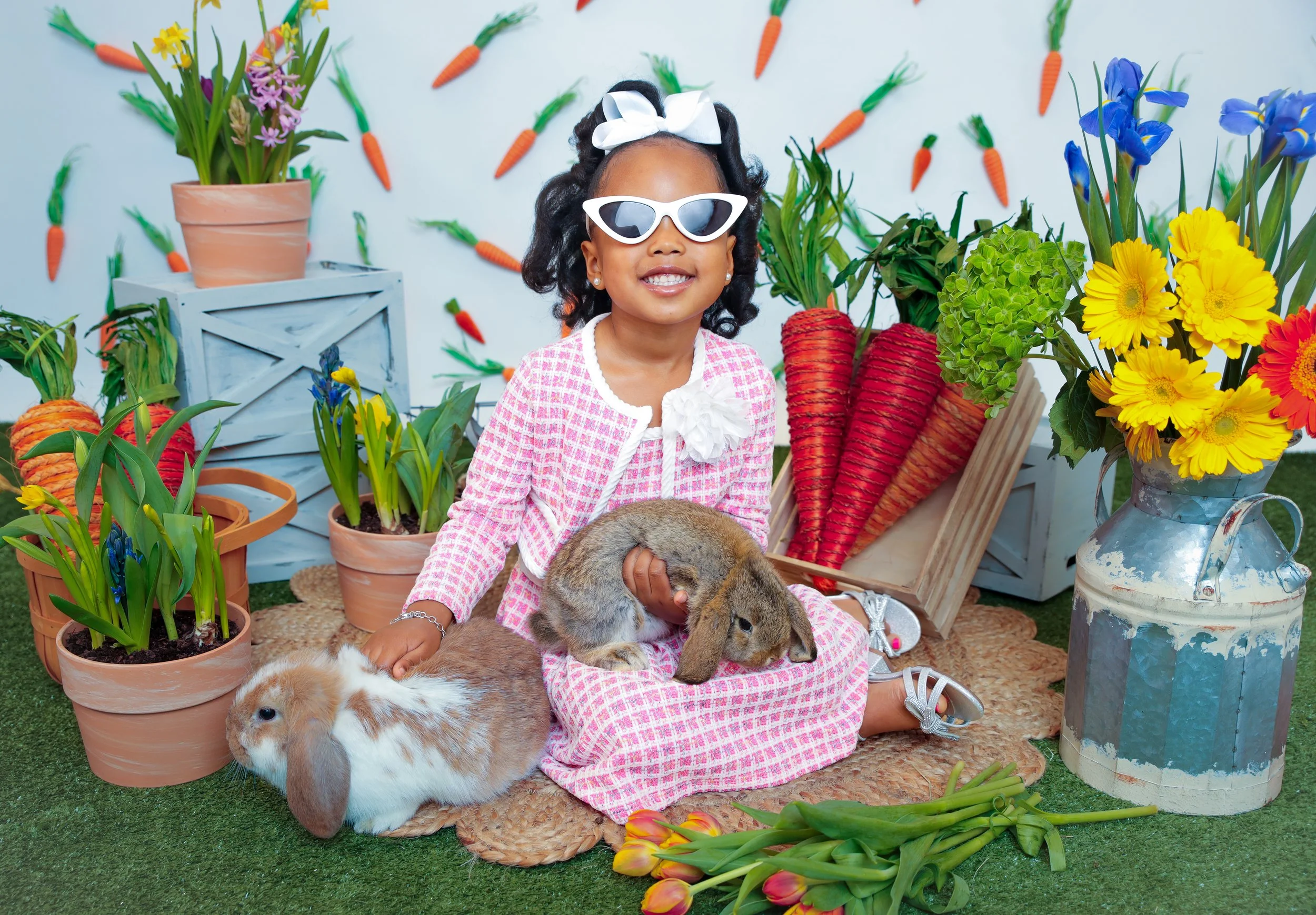 Young girl with sunglasses and white bow, sitting on a mat surrounded by potted plants and flowers, holding two rabbits, with a backdrop featuring carrot decorations.
