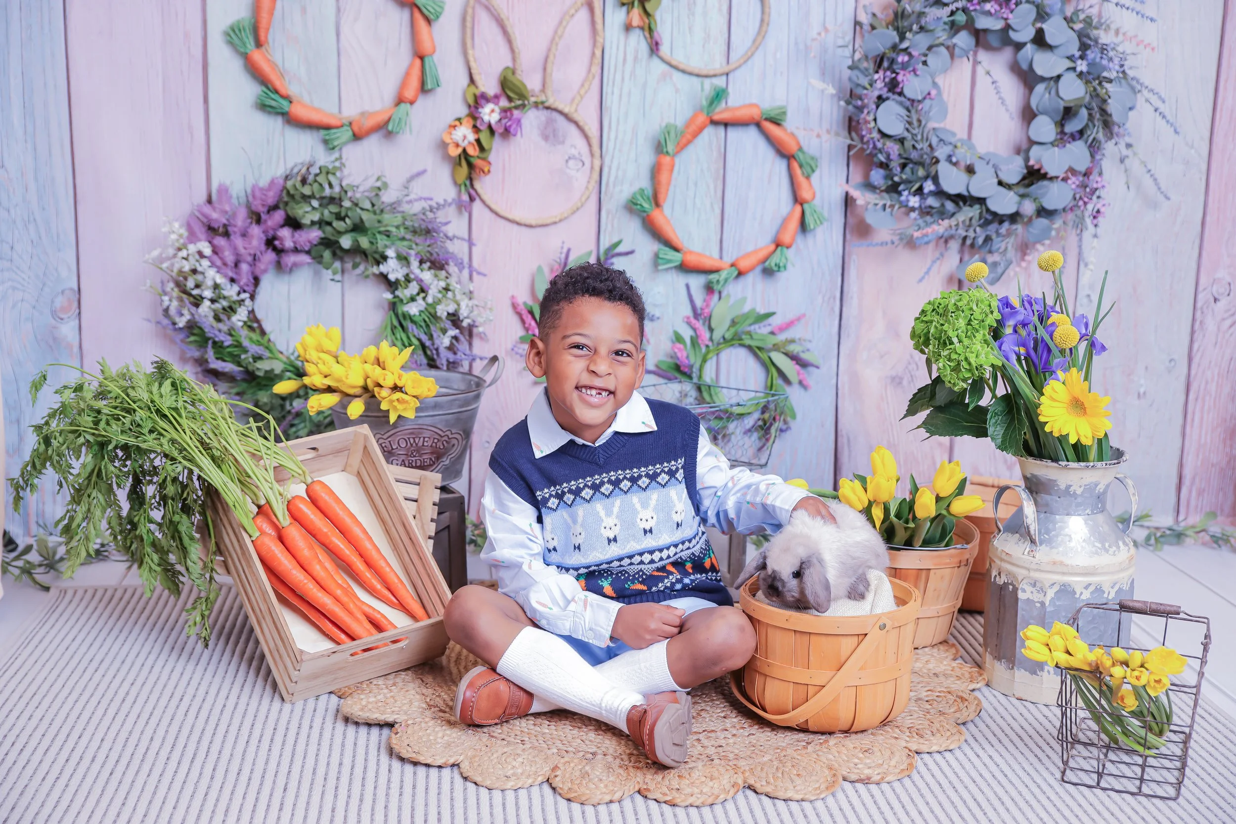 A young boy sitting on a rug with a bunny in a basket, surrounded by springtime flowers and wreaths on a pastel wooden backdrop.