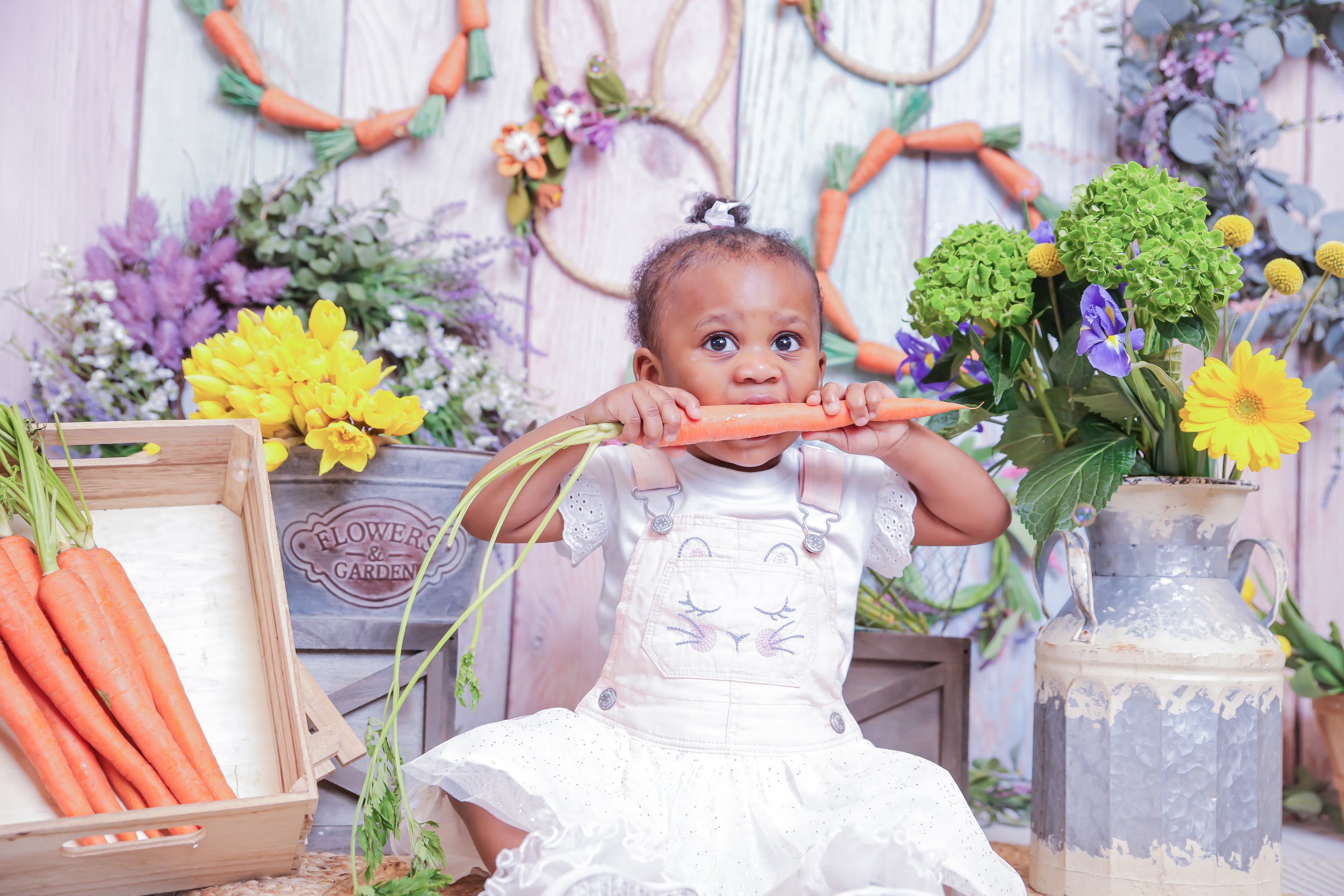 A young girl holding a large carrot in her mouth, surrounded by colorful flowers and gardening items.
