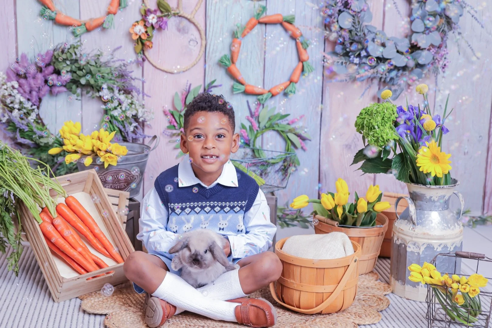 Young boy sitting cross-legged on a rug, holding a rabbit, surrounded by colorful flowers, carrots, and gardening decor.