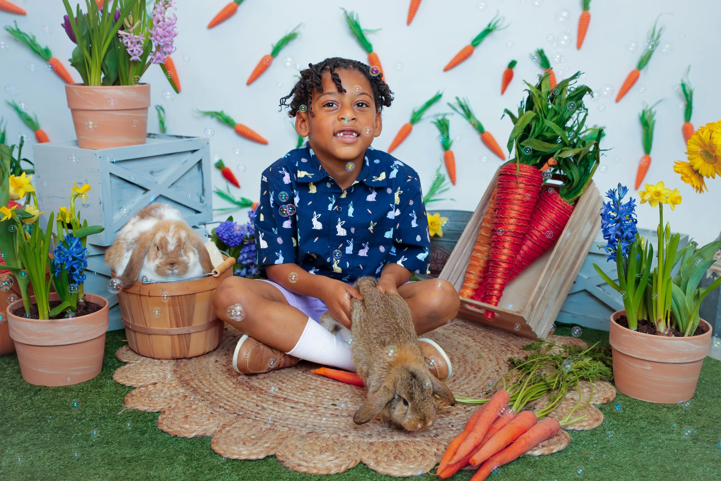 Young boy with dark curly hair and a blue shirt with rabbit patterns sitting on a round woven mat, holding a bunny rabbit. Surrounding him are potted plants, including daffodils and hyacinths, and children’s garden-themed decorations like large carro