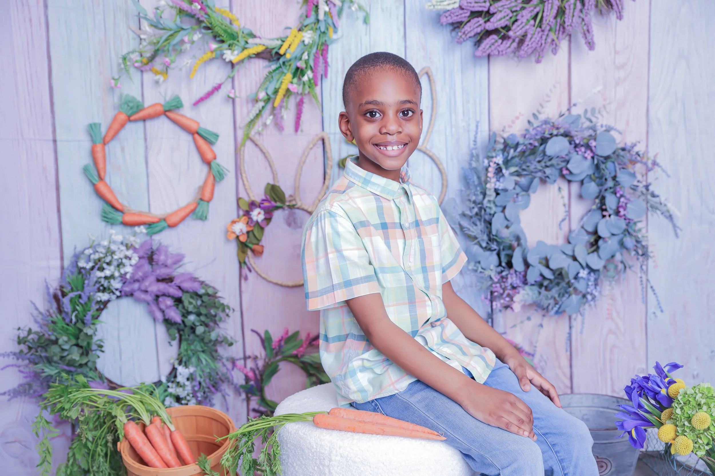 A young boy sitting on a white ottoman, smiling, surrounded by colorful floral wreaths and carrot decorations in a flower shop or garden-themed setting.