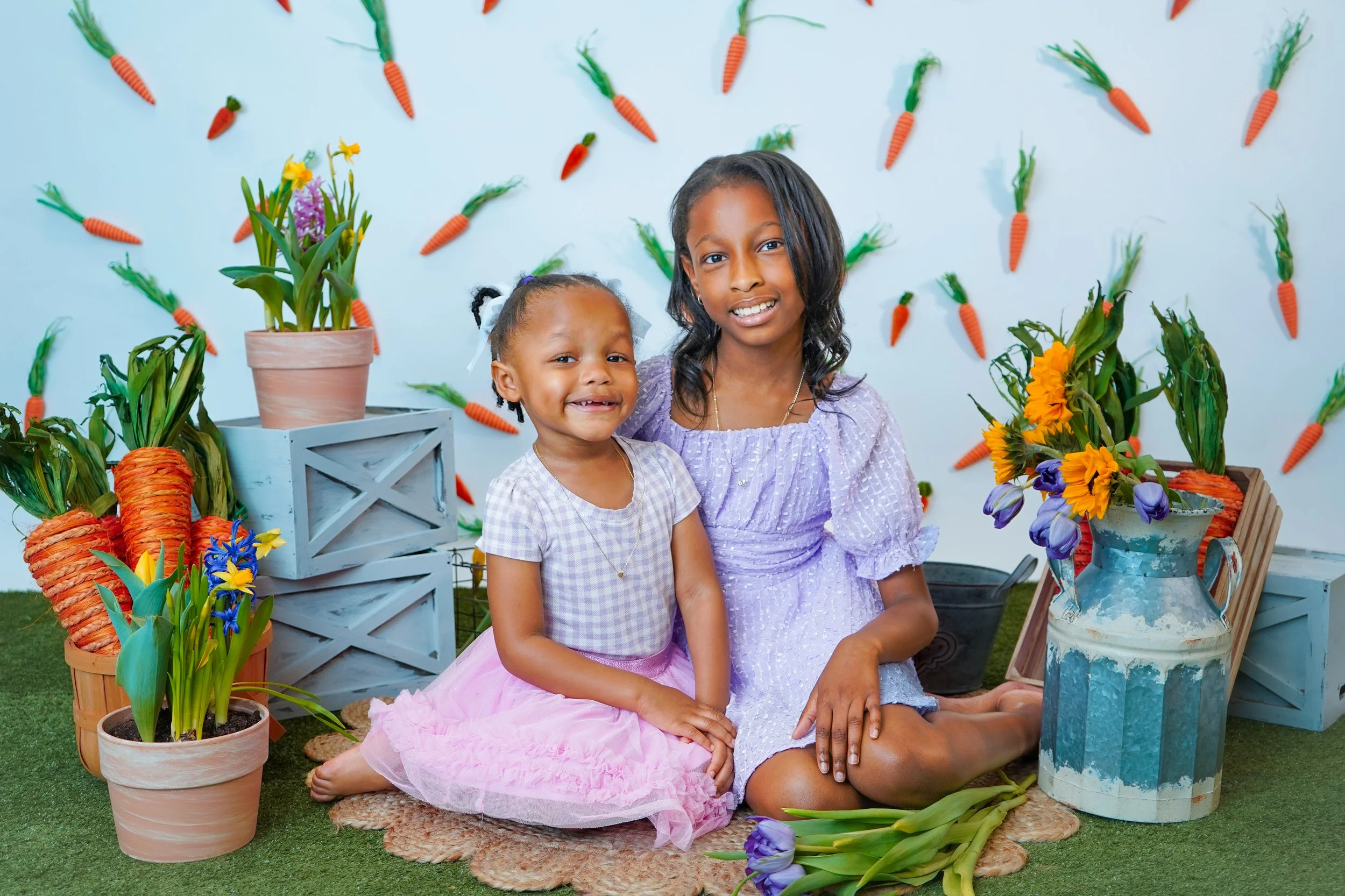 Two girls sitting on a green carpet with potted plants and sunflower decorations around them. Behind them is a wall decorated with small hanging carrots. The older girl wears a light purple dress, and the younger girl wears a light purple checkered t