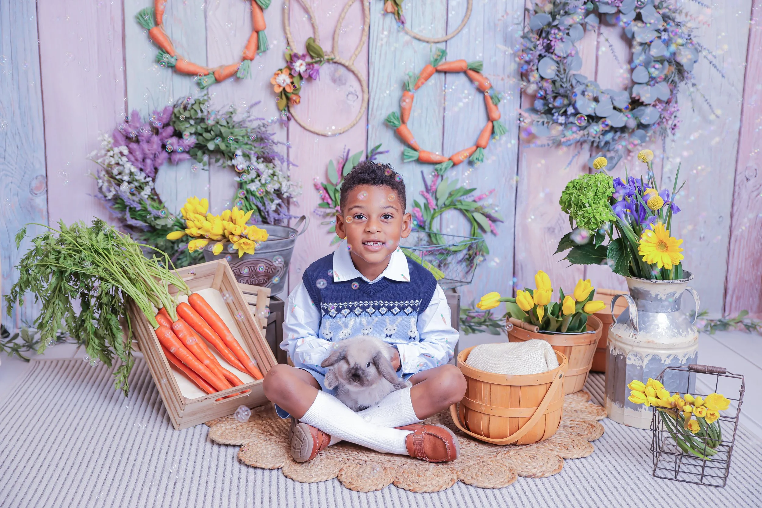 A young boy sitting cross-legged on a woven mat with a bunny in his lap, surrounded by potted flowers, carrots, and garden decor, with a pink-washed wooden wall and floral wreaths hanging behind him.