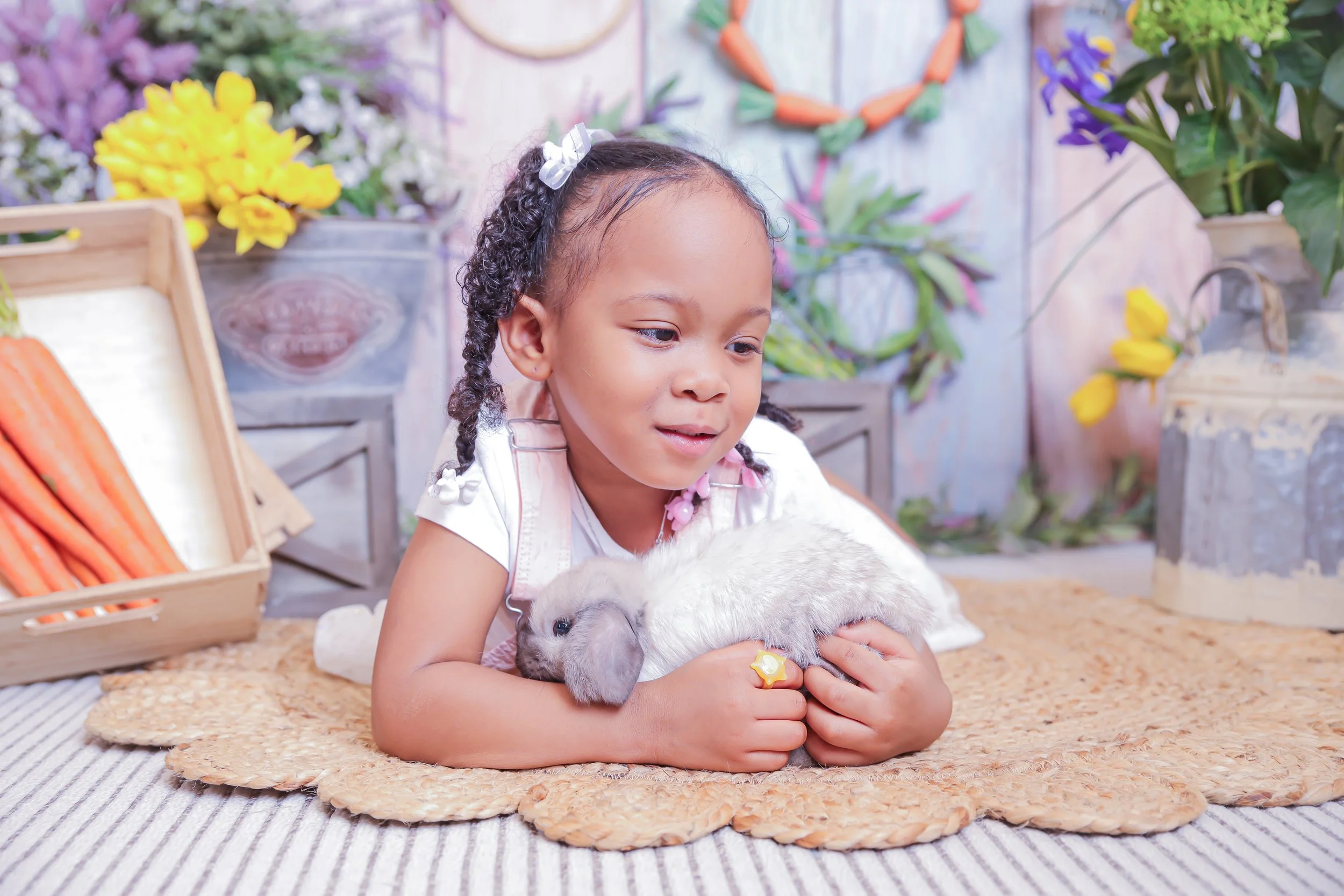A young girl with curly hair and a white bow, wearing a white shirt and pink overalls, is lying on a woven mat indoors, holding a white and gray baby rabbit. The background features colorful flowers, carrots, and decorative plants.