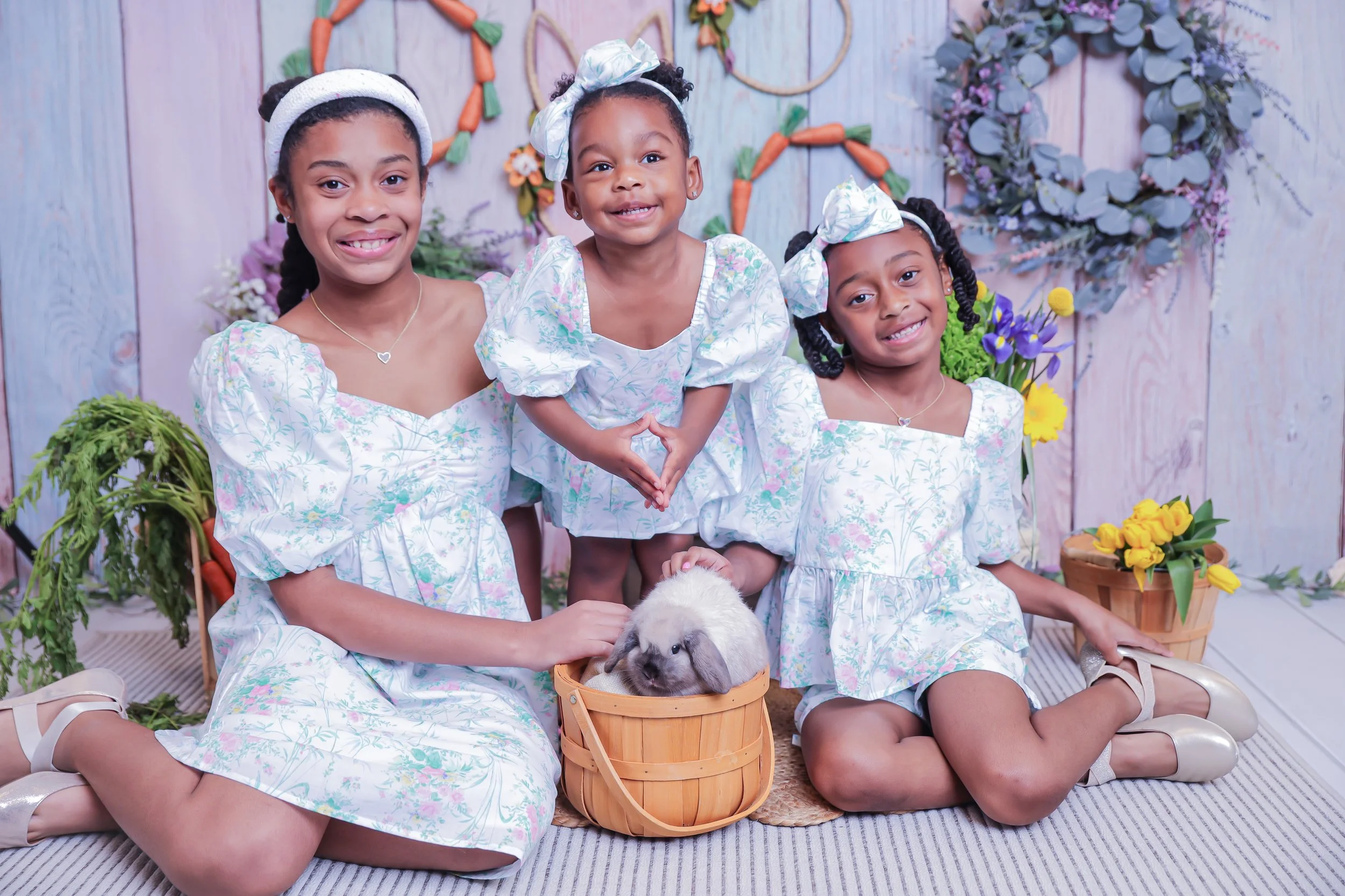 Group of four young girls with curly hair and matching white floral dresses, wearing matching headbands and bows, gathered around a rabbit in a basket, in a colorful garden setting with flowers and decorative wreaths on a wooden wall background.
