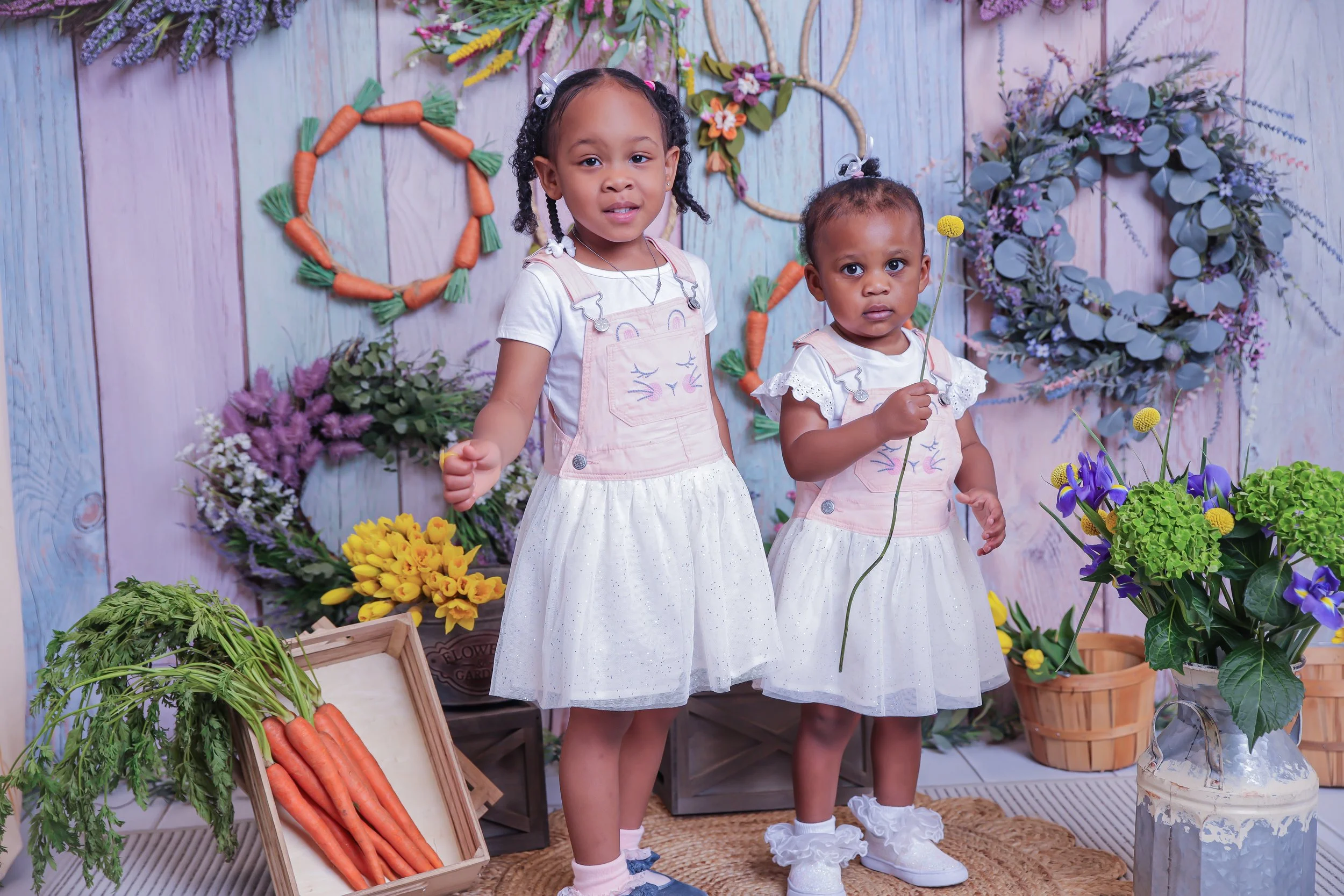 Two young girls dressed in white dresses with pink overalls, standing in front of a pastel-colored wooden wall decorated with floral wreaths and carrot-shaped decorations. The girls are holding flowers, with one holding a yellow flower and the other 