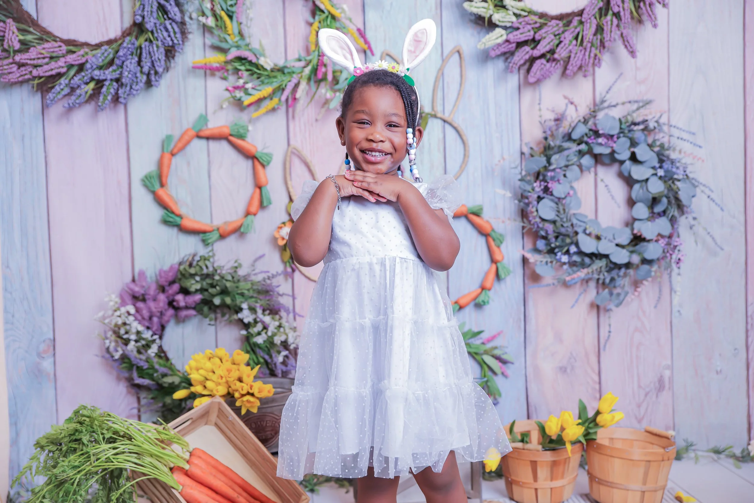 A young girl dressed in a white dress with bunny ears headband, smiling with her hands on her chin, standing in front of a spring-themed backdrop with floral wreaths, baskets of carrots and yellow flowers.
