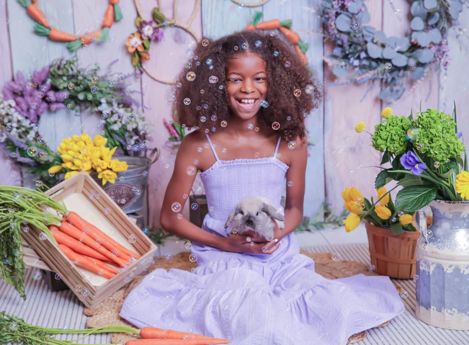 A girl with curly hair wearing a white dress, smiling, holding a small bunny, surrounded by colorful flowers, carrots, and garden props, in a vibrant floral setting.