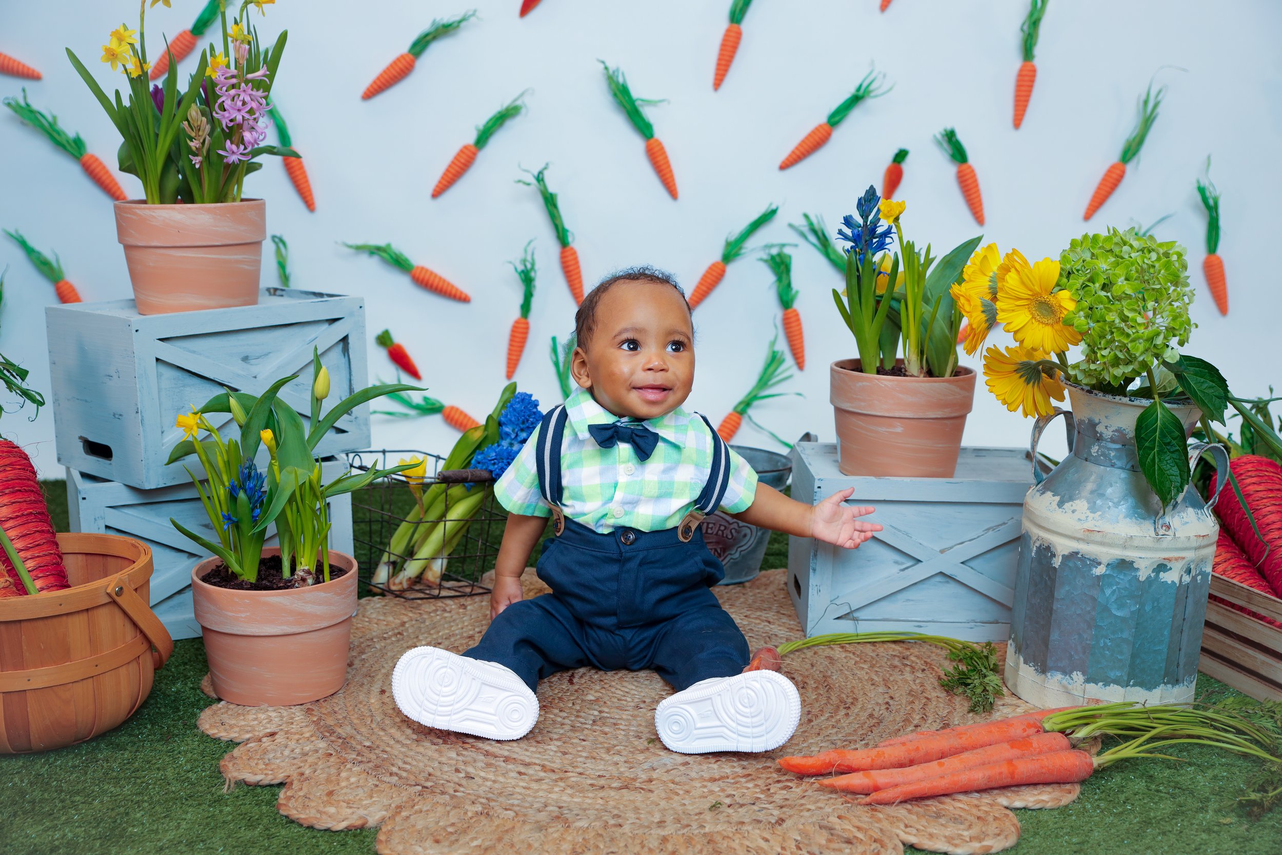 A smiling baby boy sitting on a woven mat surrounded by potted flowers, carrots, and decorative garden elements, with a wall of carrot decorations in the background.