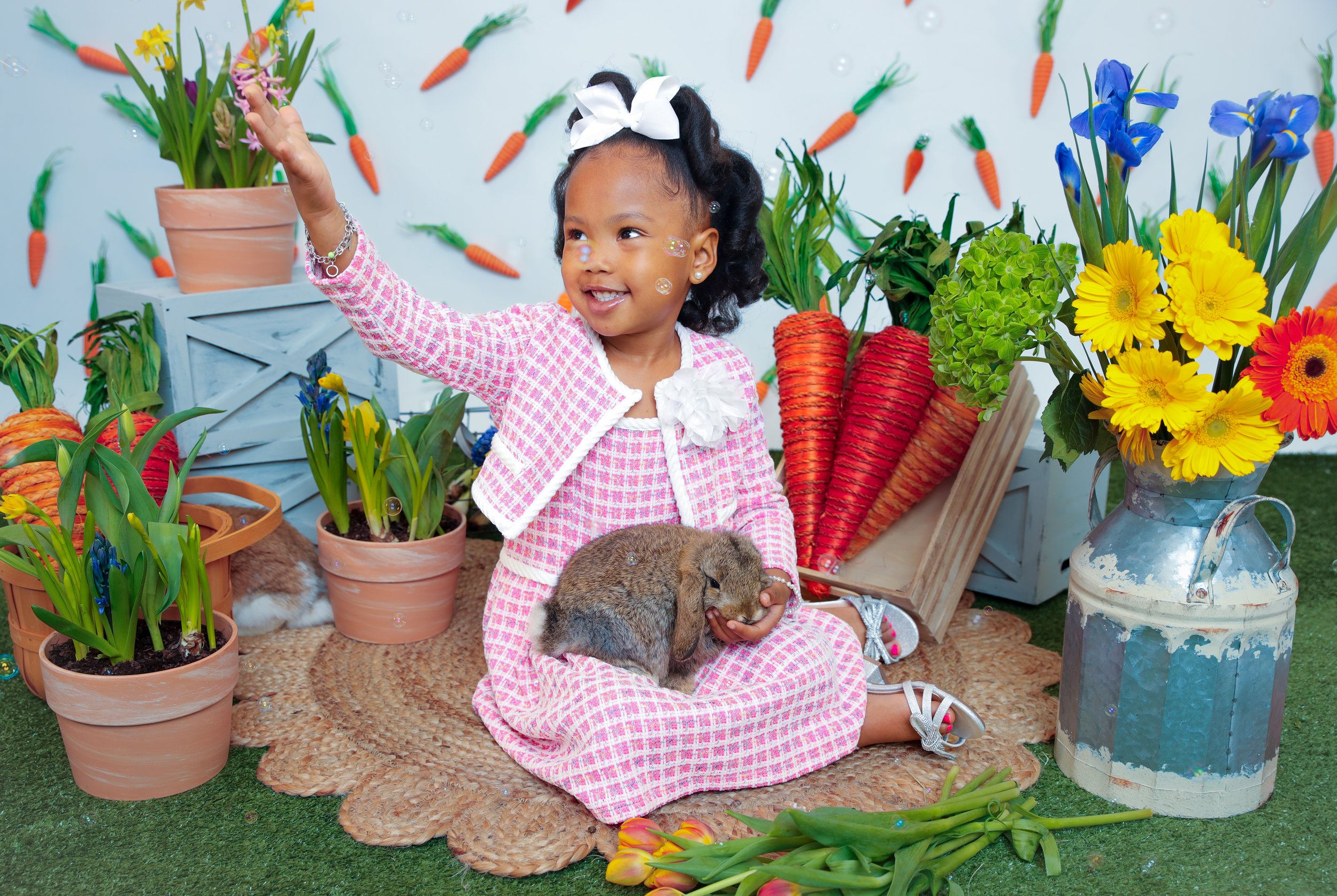 A young girl sitting on a woven mat surrounded by potted tulips, daffodils, and decorative carrots. She's holding a small bunny and smiling, with bubbles floating around her.