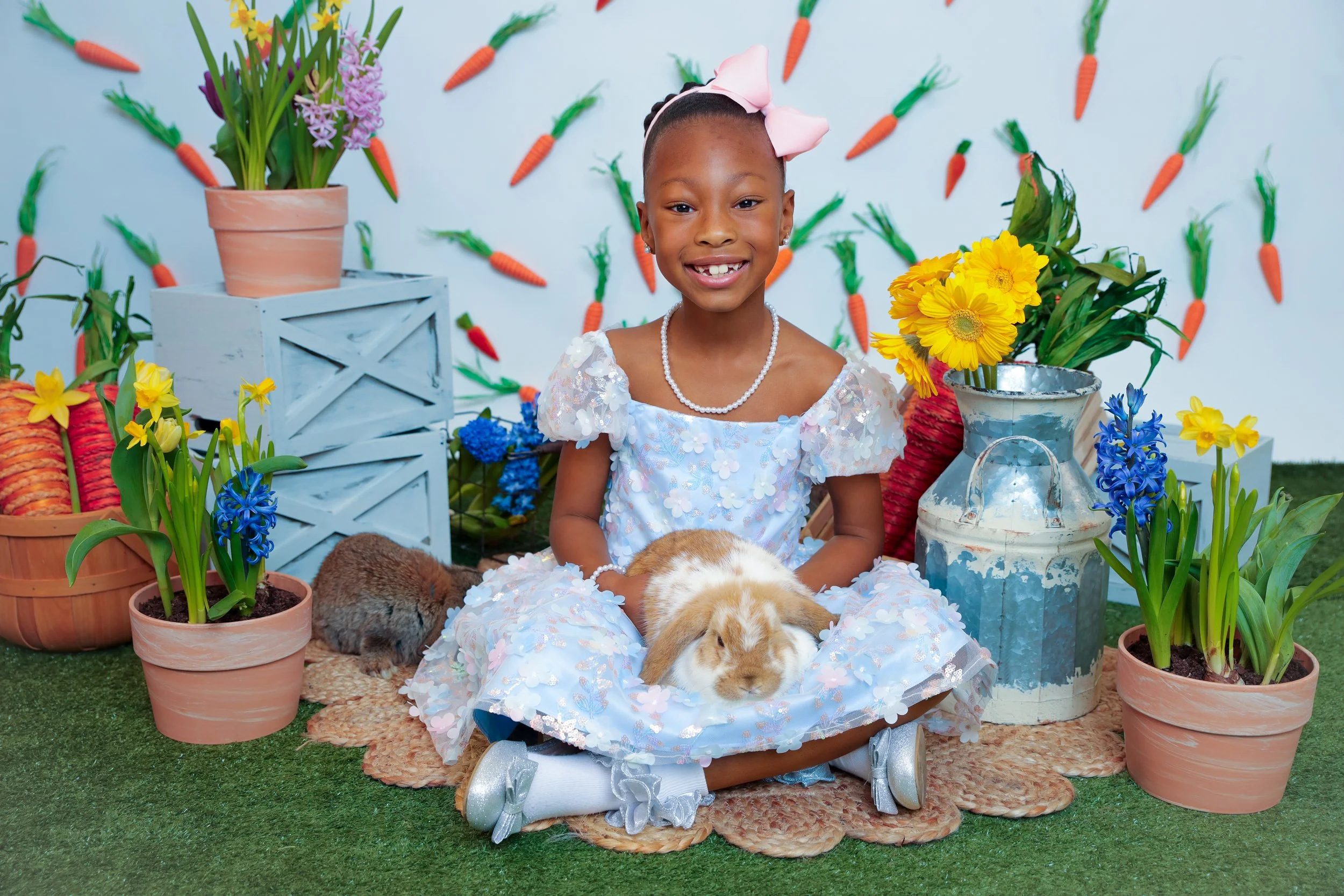 A young girl with a pink bow in her hair, wearing a white dress with floral embellishments and a pearl necklace, sits on a woven rug holding a brown and white rabbit on her lap. The background features a white wall decorated with hanging paper carrot