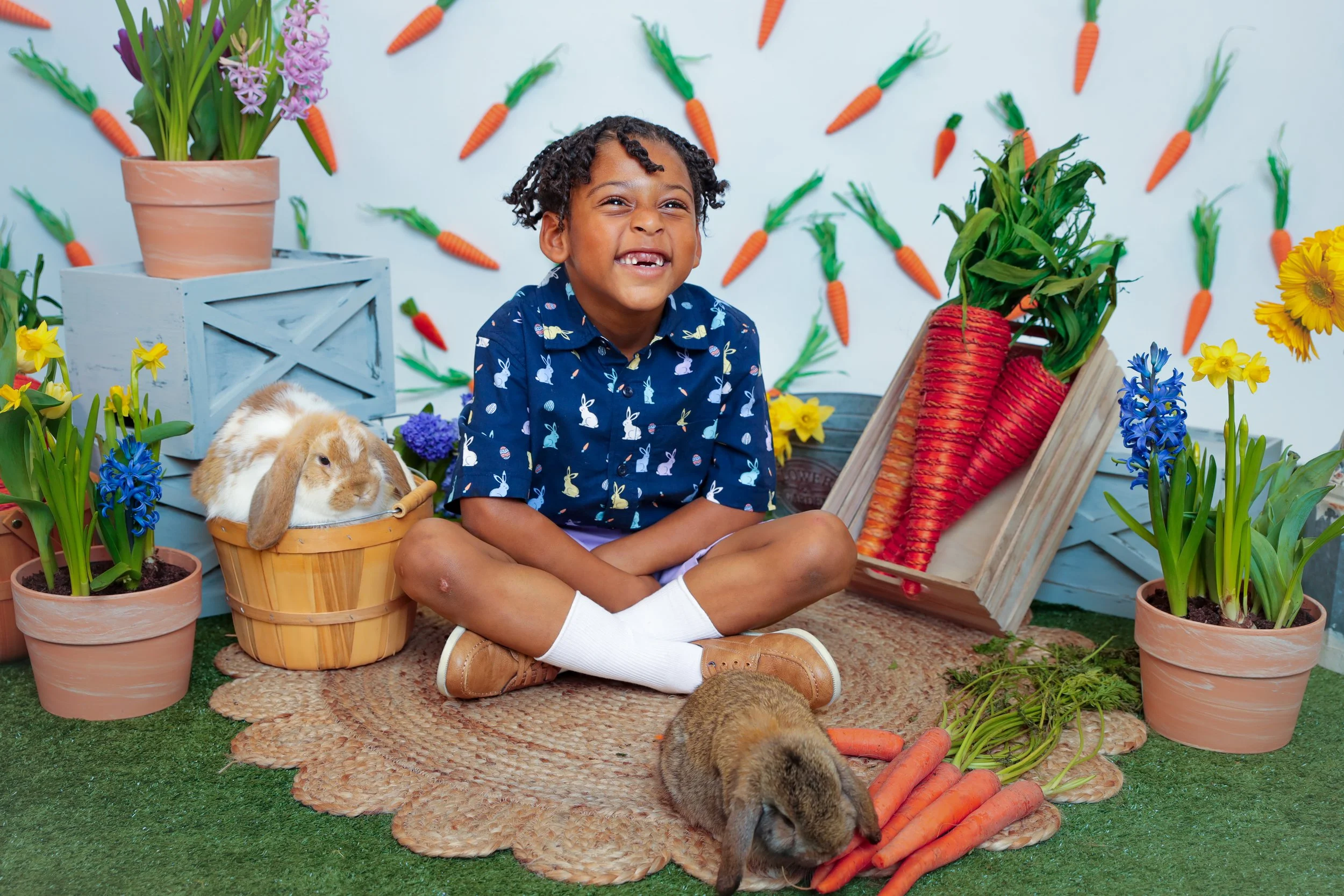 A young boy sitting cross-legged on a woven rug, smiling with excitement, surrounded by spring flowers, carrots, and bunny decorations, in an Easter-themed setting.
