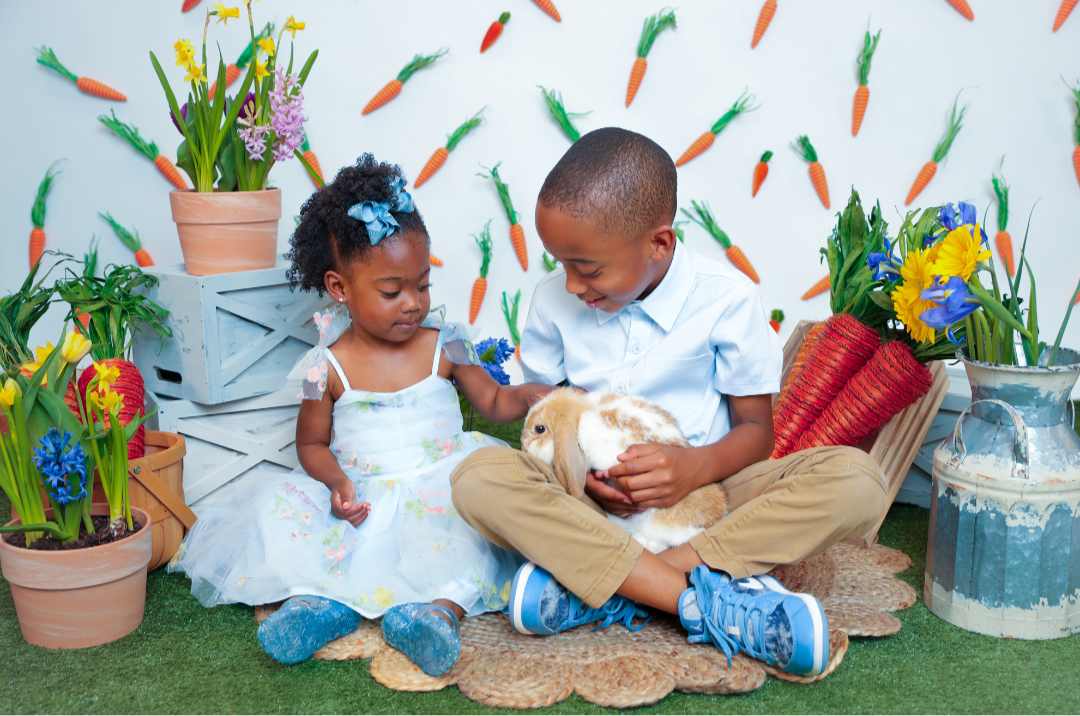 Two children, a young girl and a boy, sitting on a rug outdoors, holding a rabbit, surrounded by potted flowers and a backdrop decorated with carrots.