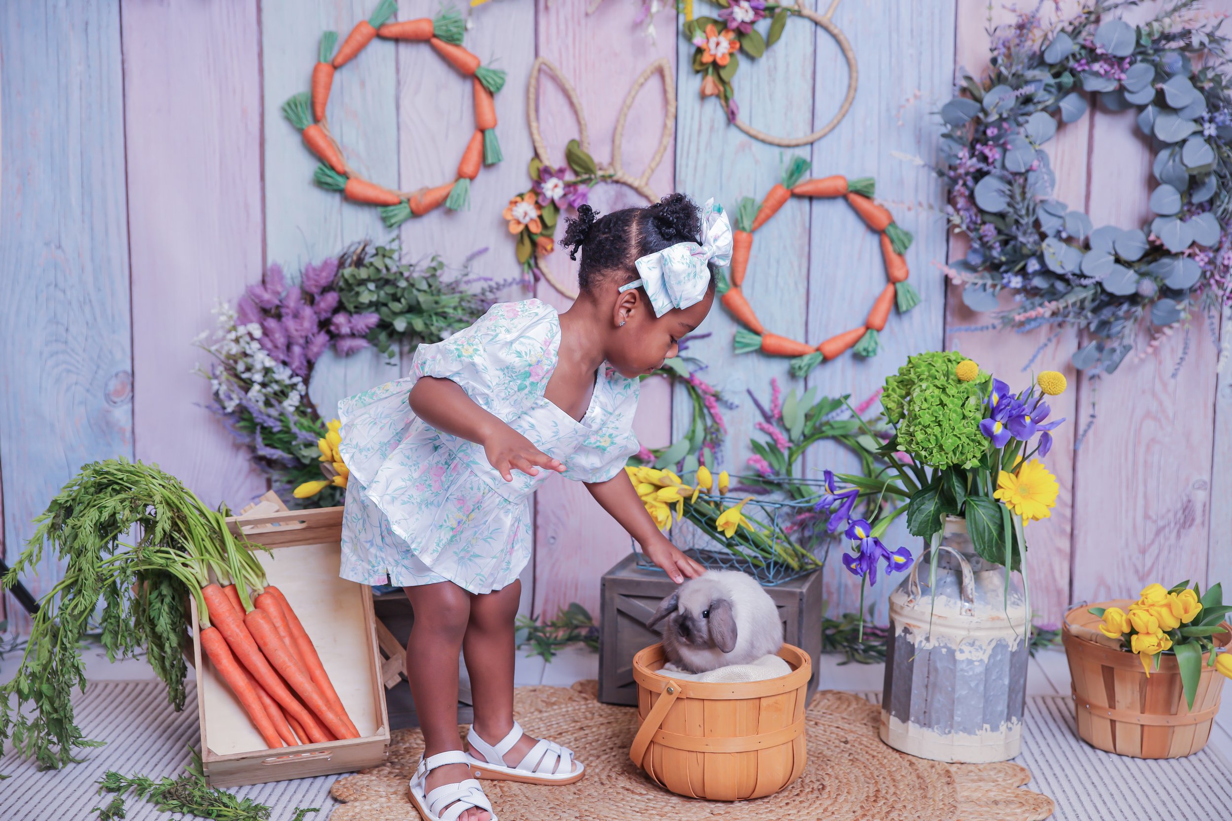 A young girl with a white dress and matching bow petting a gray bunny inside a wooden basket, surrounded by colorful flowers and decorative vegetable wreaths on a pastel-colored wooden wall background.