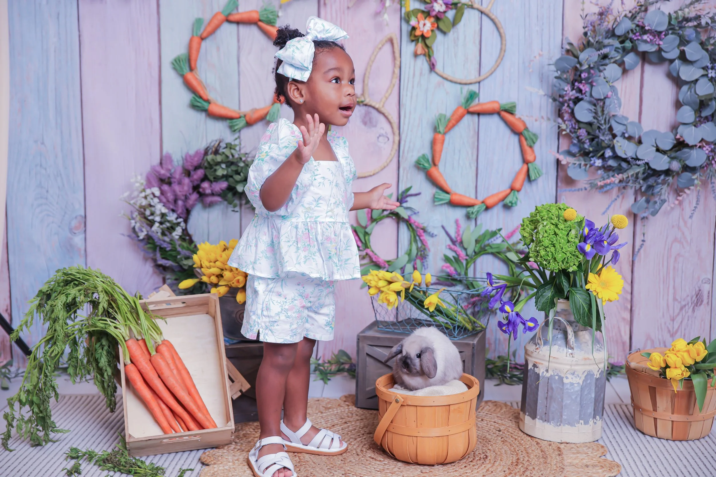 A young girl dressed in light blue floral pajamas stands with her hands raised as if talking or explaining, in a colorful setting with a backdrop of pastel-colored wooden panels. The scene is decorated with vibrant flowers, including purple, yellow, 