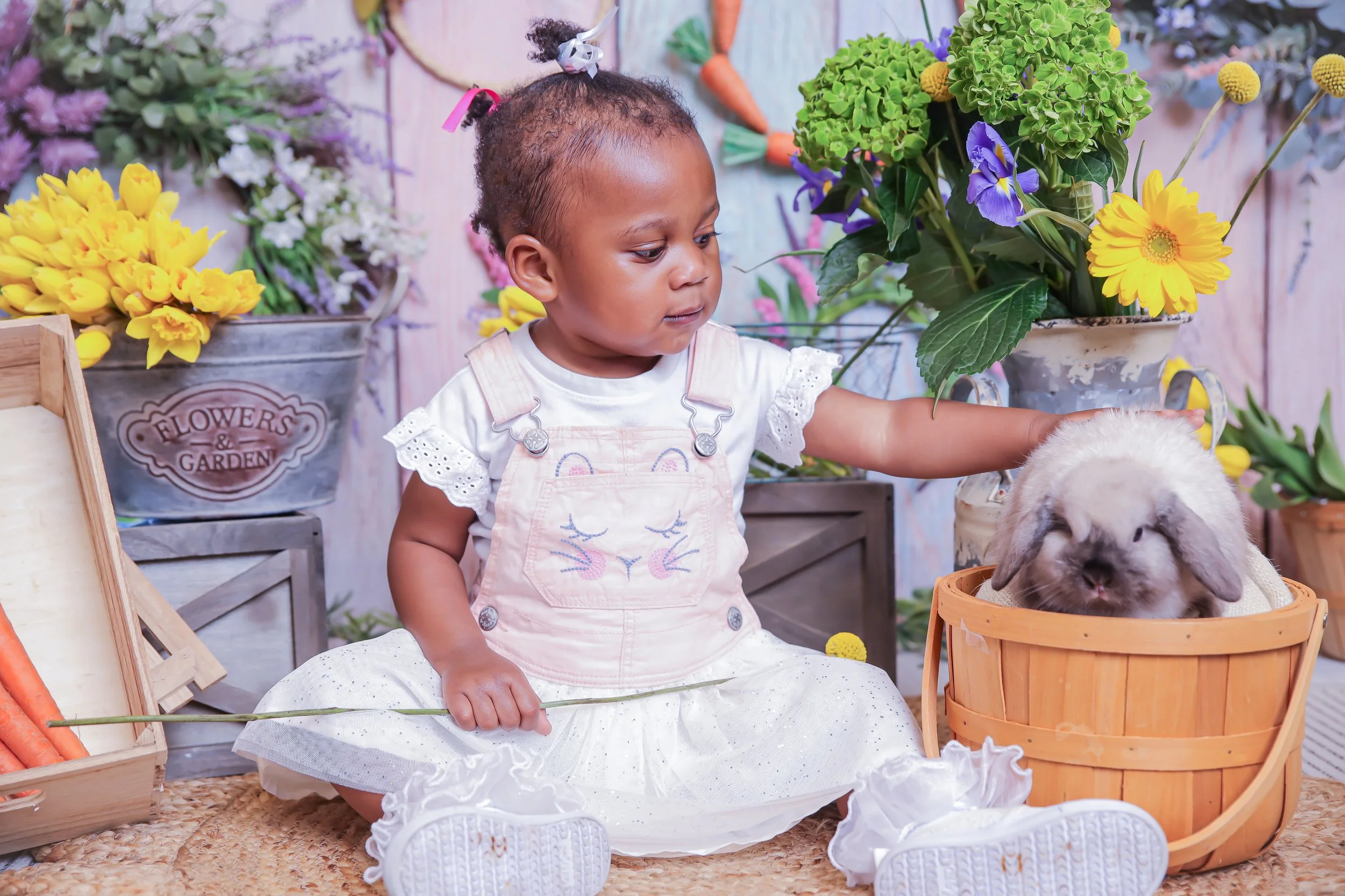 A young girl in a white dress with eyelet lace and bunny embroidery, sitting on a woven mat, petting a fluffy white rabbit inside a wooden basket, surrounded by flowers and gardening props.