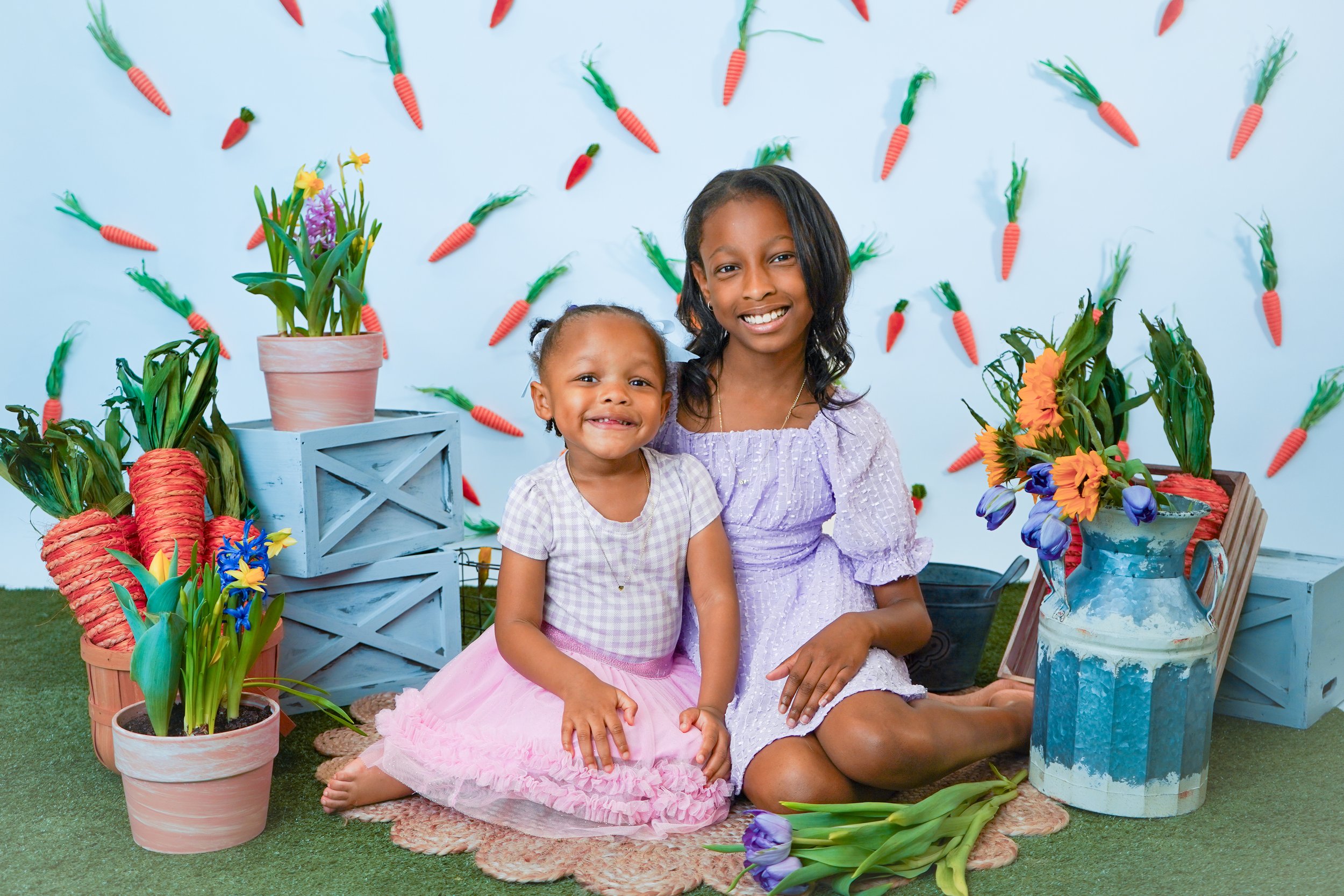 A woman and a young girl sitting on a blanket surrounded by flowers and spring-themed decorations in a spring or Easter celebration setting.