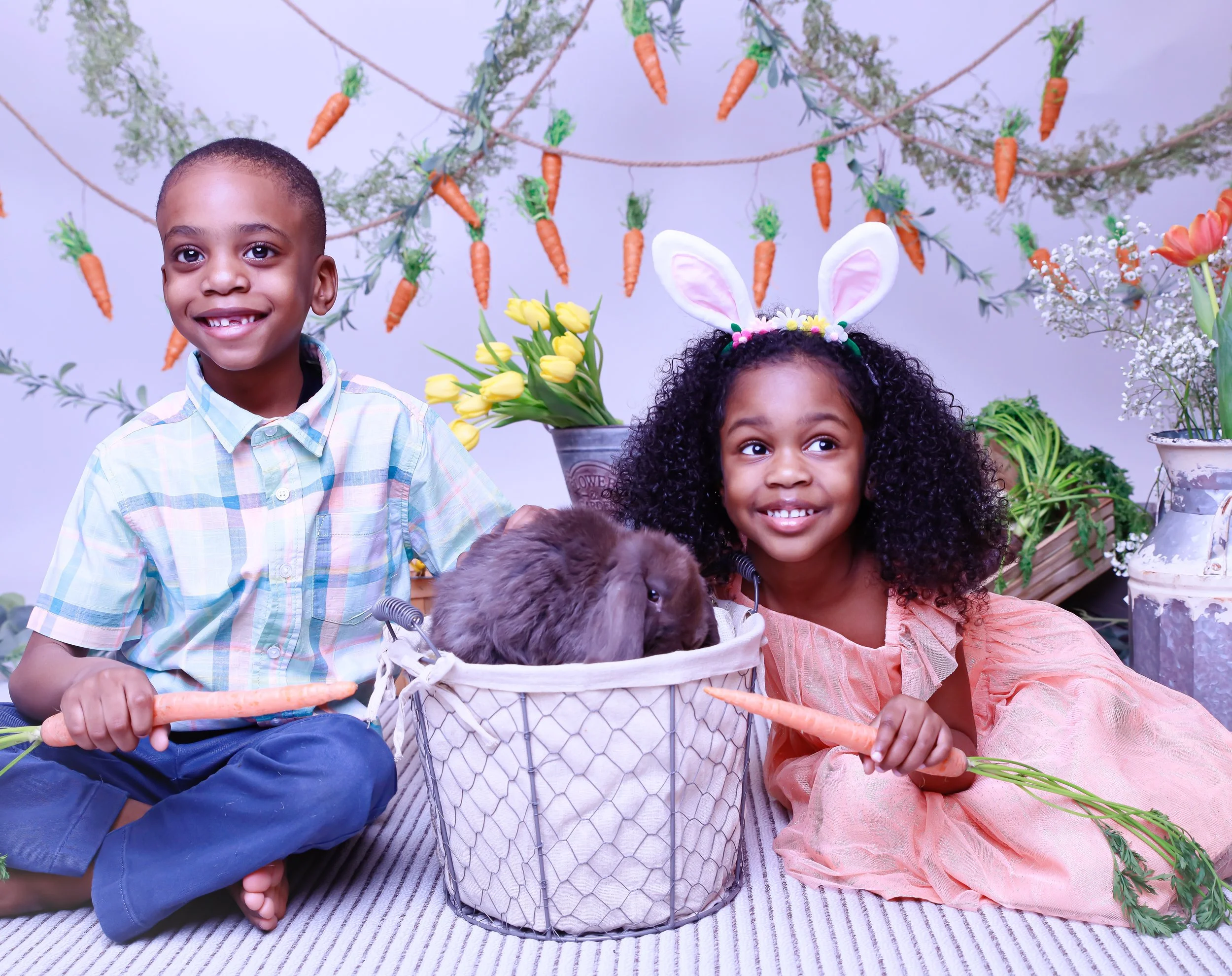 Two children, a boy and a girl, with a rabbit in a basket, holding carrots, with flowers and carrots hanging in the background, celebrating Easter.