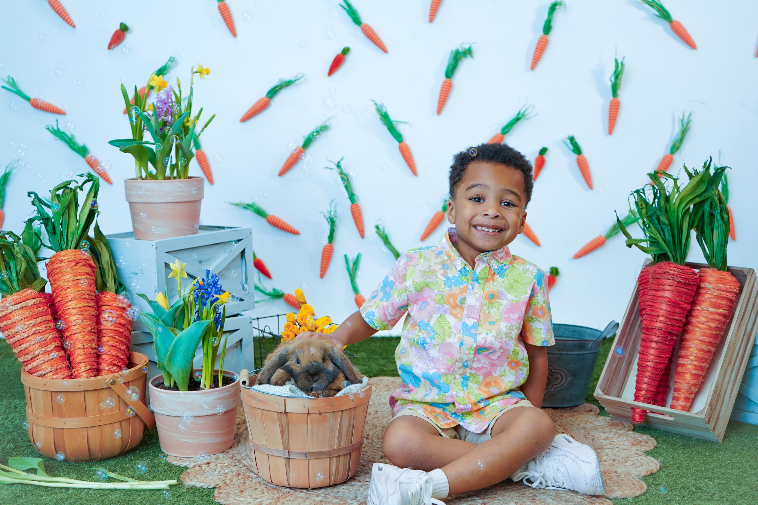A young boy sitting on a rug with a rabbit in a basket, surrounded by carrots, potted flowers, and decorative carrots on a white backdrop.