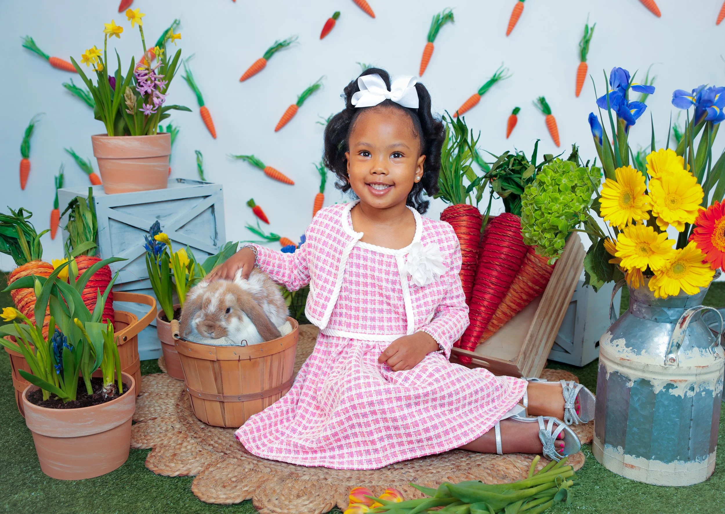 A young girl in a pink dress and white cardigan sitting on a woven mat surrounded by spring flowers, big carrots, and a bunny rabbit in a basket with a colorful, festive background.