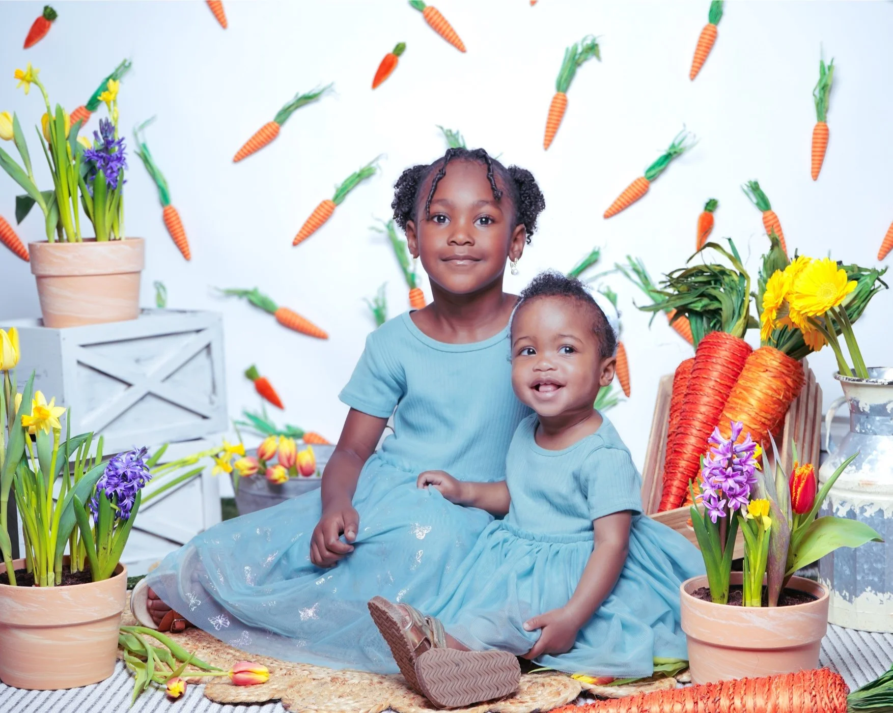 Two young girls wearing light blue dresses sitting on a woven rug surrounded by potted flowers and large carrots as decorations. The background is decorated with hanging artificial carrots and more flowers.