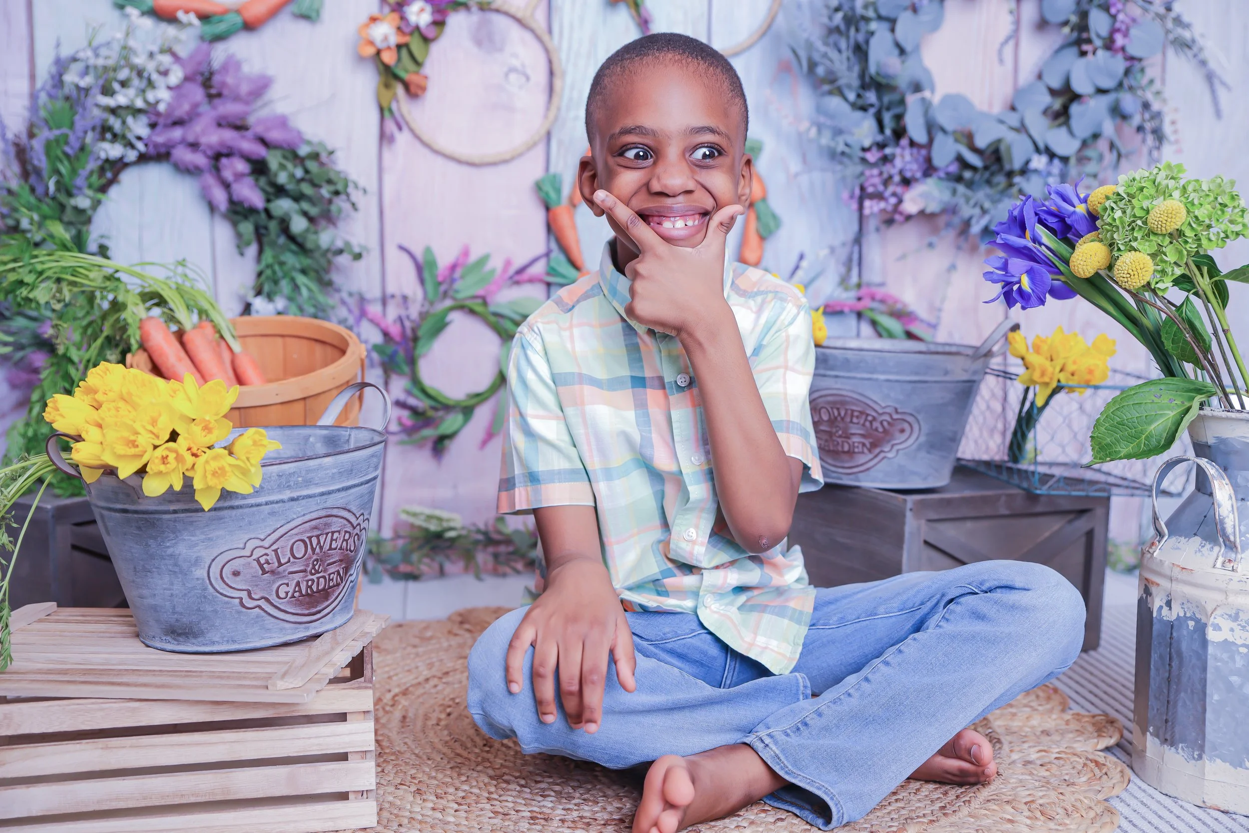 Child sitting cross-legged on a woven mat, smiling and making a funny face, surrounded by colorful flowers and gardening supplies in a garden-themed setting.