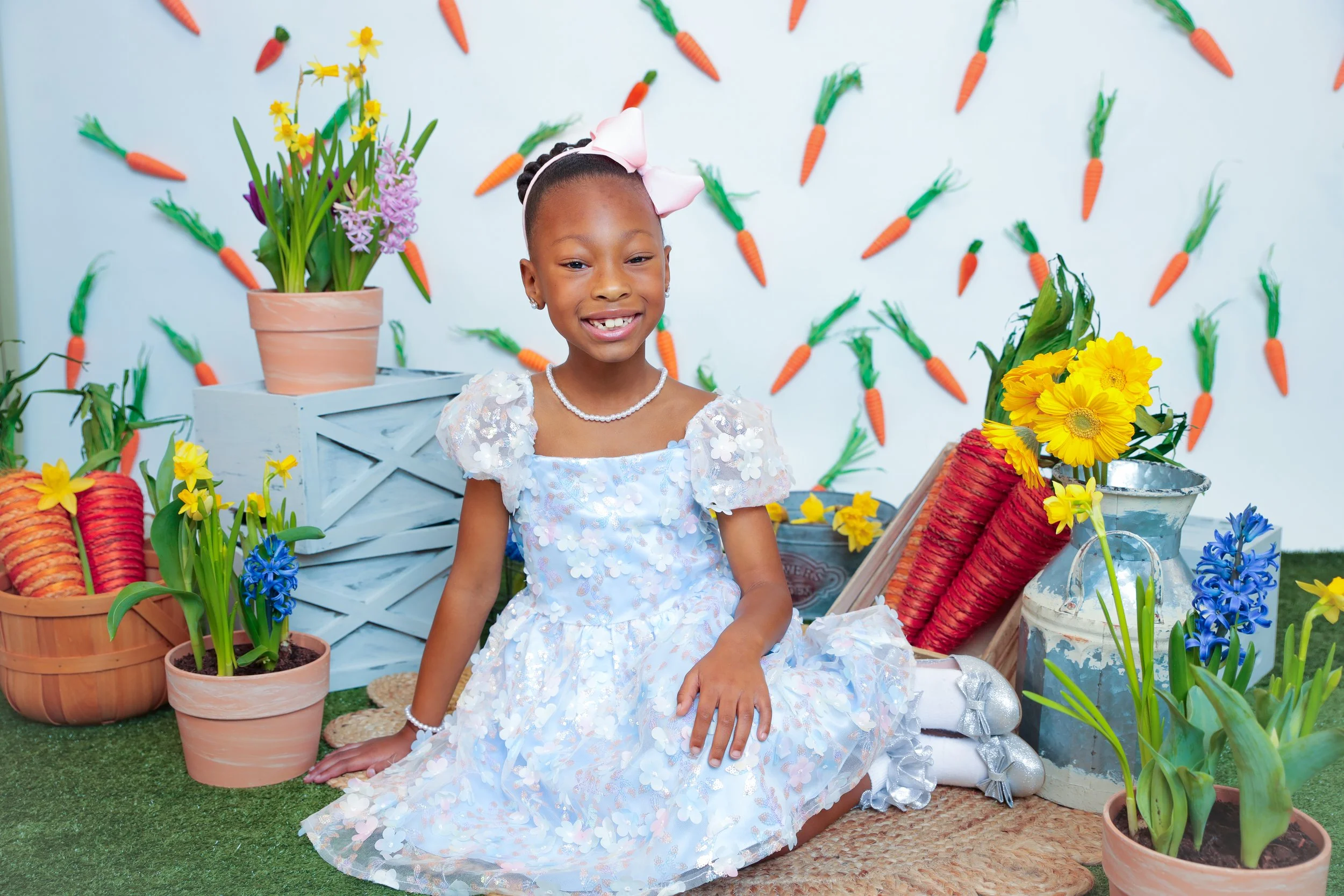 A young girl in a white dress sitting on a green grass mat surrounded by potted flowers and paper carrots, with a white wall decorated with paper carrots in the background.
