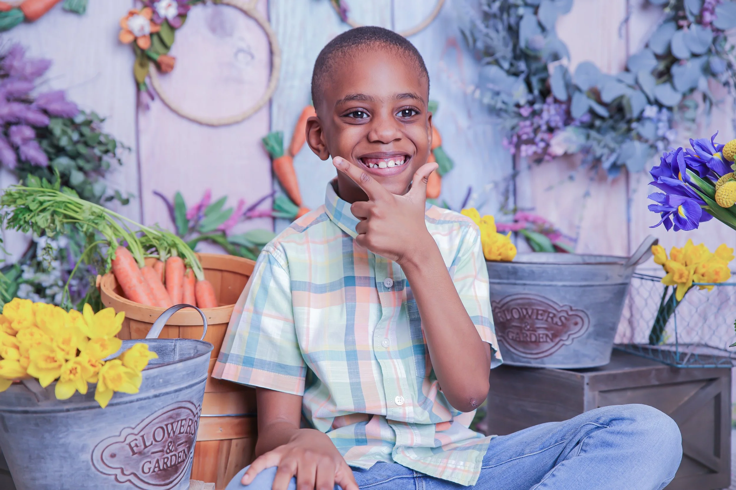 A young boy sitting on a bench at a flower and vegetable market, smiling and making a peace sign with his fingers. There are baskets of carrots and flowers around him.