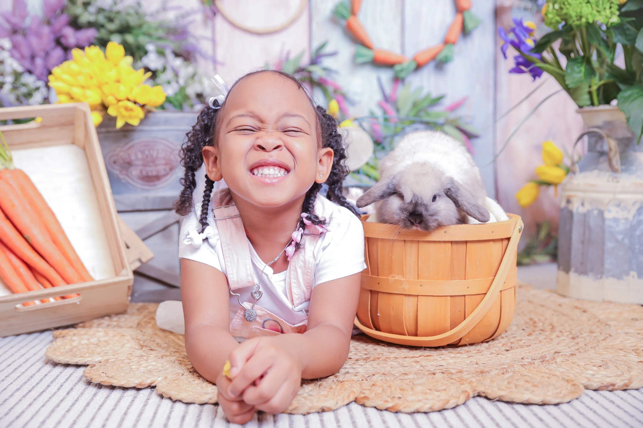 A young girl with curly hair smiling happily, lying on her stomach on a woven mat, with a small grey and white rabbit sitting in a wooden basket next to her. The background is decorated with colorful flowers and vegetable props.