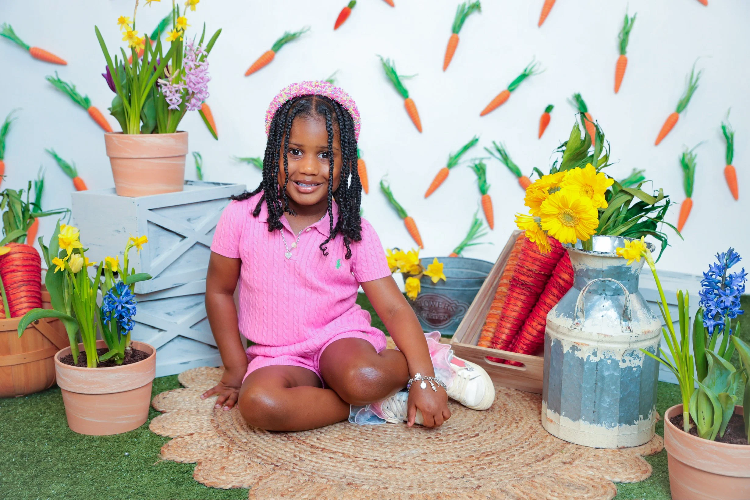 A young girl with braids and a pink outfit sitting on a woven rug among potted flowers and decorative items, with a backdrop decorated with orange carrots and green foliage.