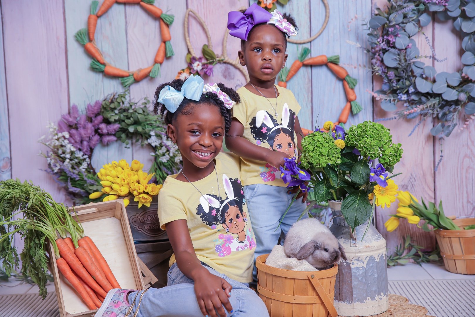 Two young girls with bows in their hair, wearing matching yellow T-shirts with a bunny girl graphic, cheerful expressions, sitting among flowers and carrots, with a stuffed bunny in a basket, set against a spring-themed decorated background.