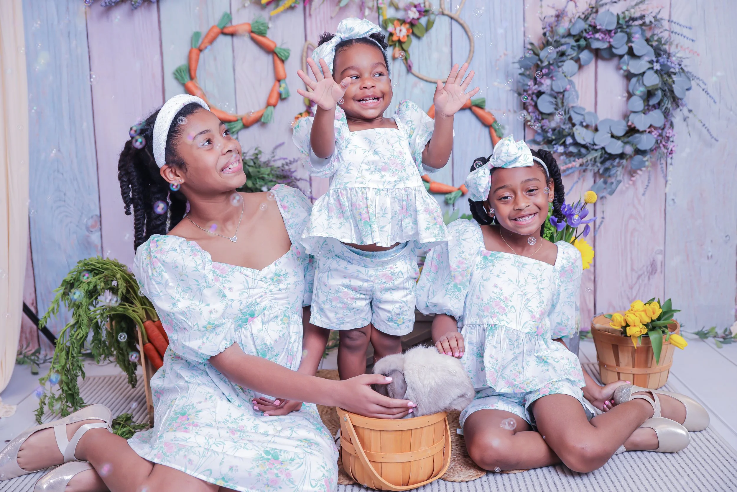 Four young girls in matching floral dresses and headbands, smiling and playing with a rabbit in a decorated indoor setting with flowers, carrots, and a pastel-colored wooden backdrop.