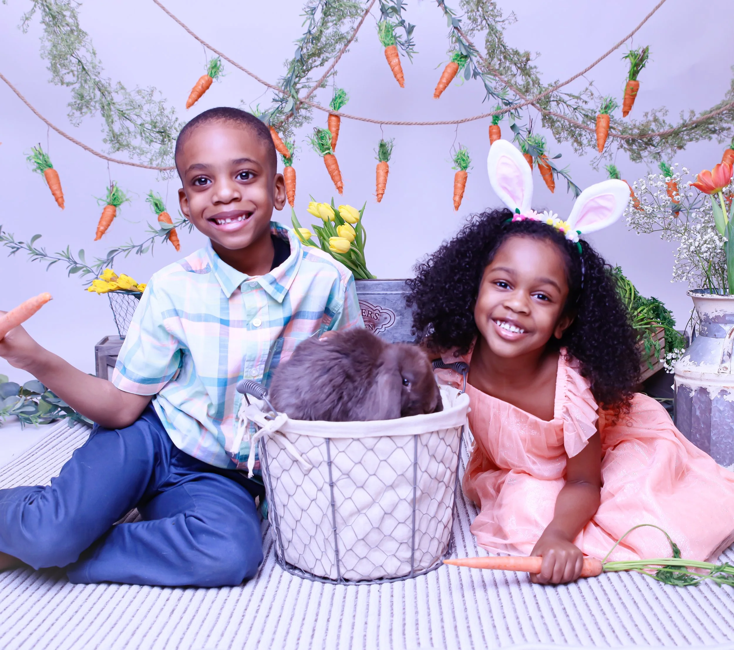 Two children, a boy and a girl, sitting on the floor with a rabbit in a basket, surrounded by springtime decorations like hanging carrots, flowers, and bunny ears, smiling at the camera.