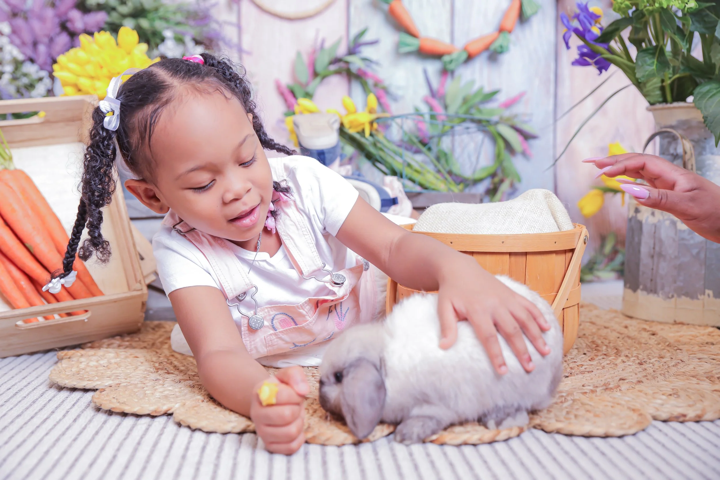 A young girl playing with a white rabbit at a flower shop or market, with carrots and colorful flowers in the background.