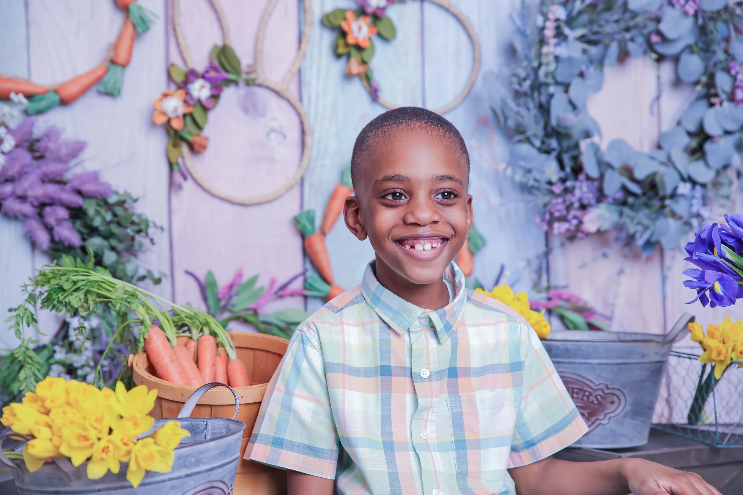 A young boy smiling in front of a colorful background with flowers and carrots, surrounded by flower buckets and decorative wreaths.