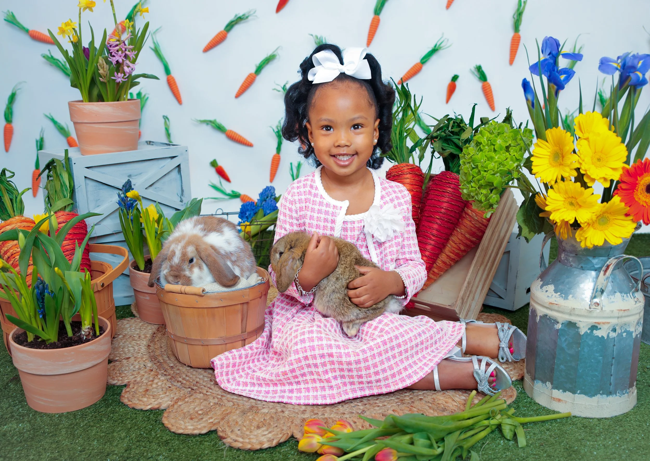 A young girl sitting on a rug, holding two bunnies, surrounded by flowers and carrots, with a backdrop of more carrots, flower arrangements, and potted plants.