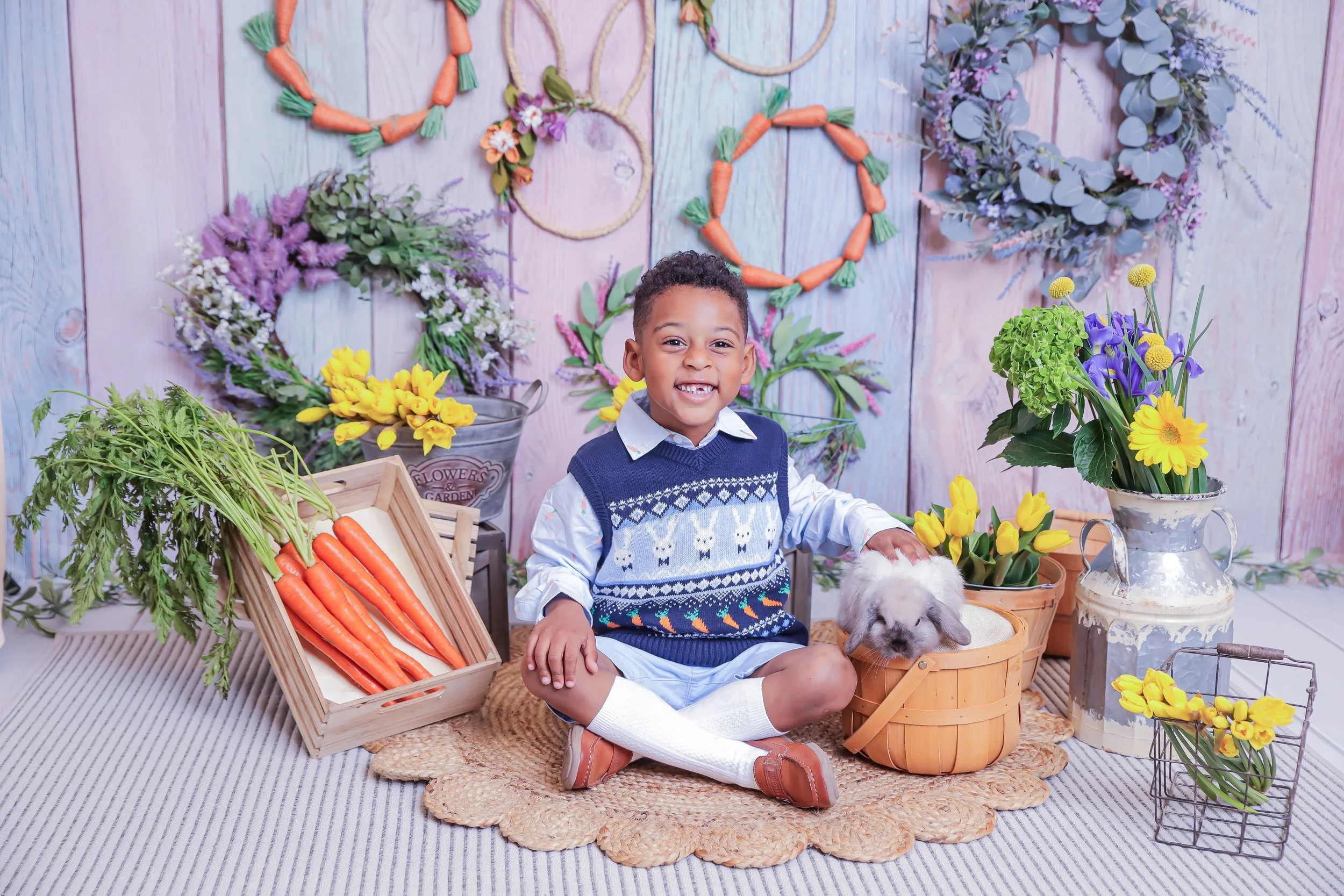A young boy sitting on a woven rug with a small bunny in a basket, surrounded by colorful flowers and decorative wreaths on a pastel wooden backdrop.