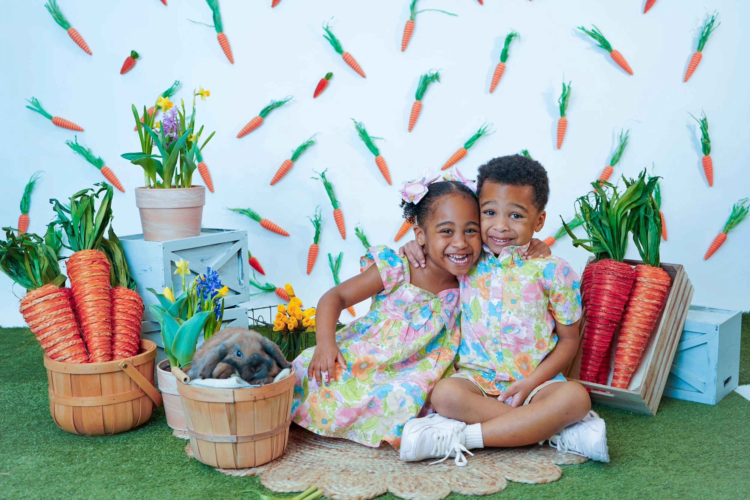 Two children hugging and smiling, surrounded by Easter-themed decorations including large paper carrots and bunny-ear headband, with flowers and bunny plush on a grassy floor.