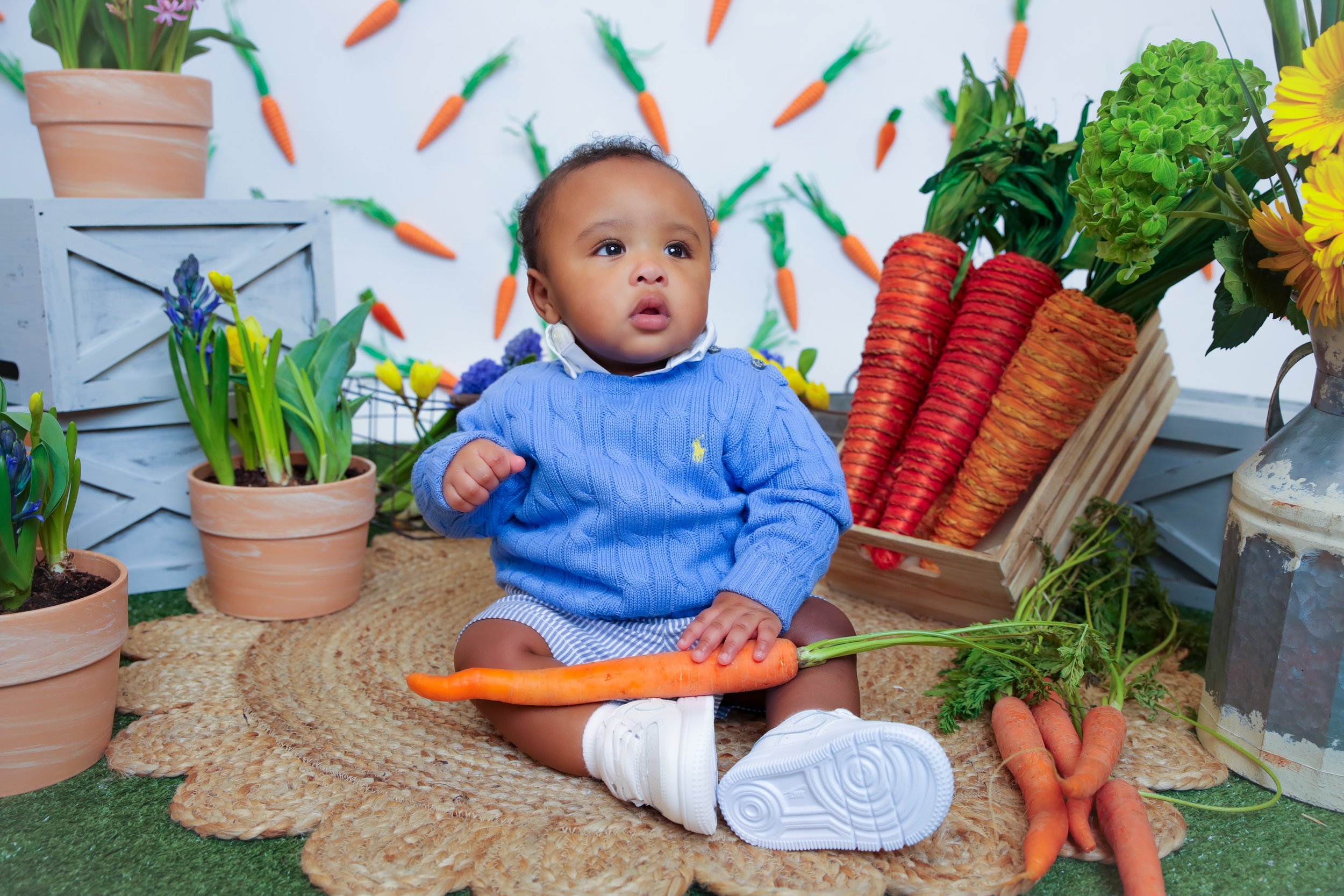 Young child sitting on a woven rug surrounded by potted plants, carrots, and decorative veggies with a backdrop of carrot decorations on the wall.